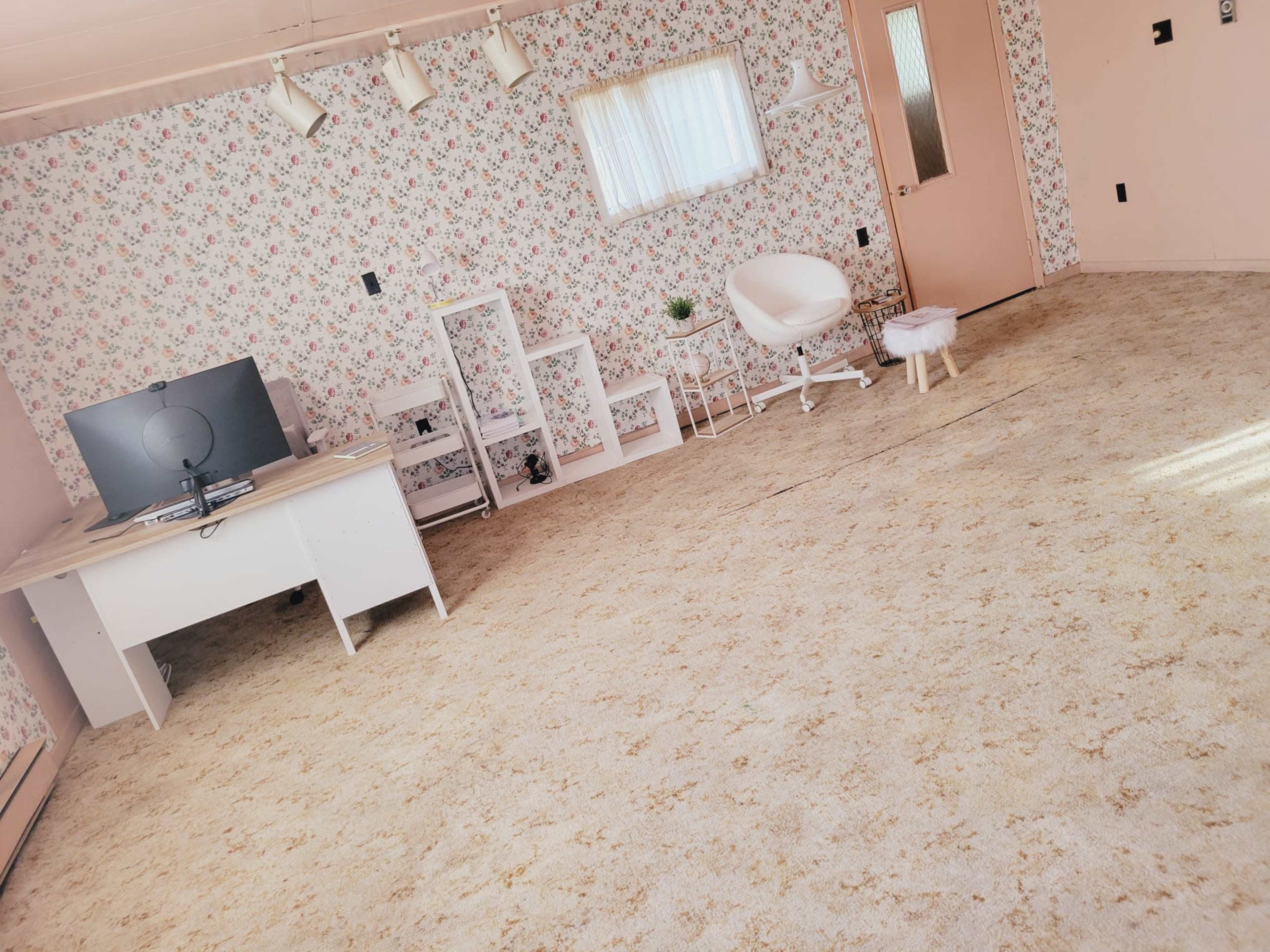 The image shows a light-colored room with floral wallpaper, a desk with a computer monitor, a small bookshelf, a potted plant, and a white stool.