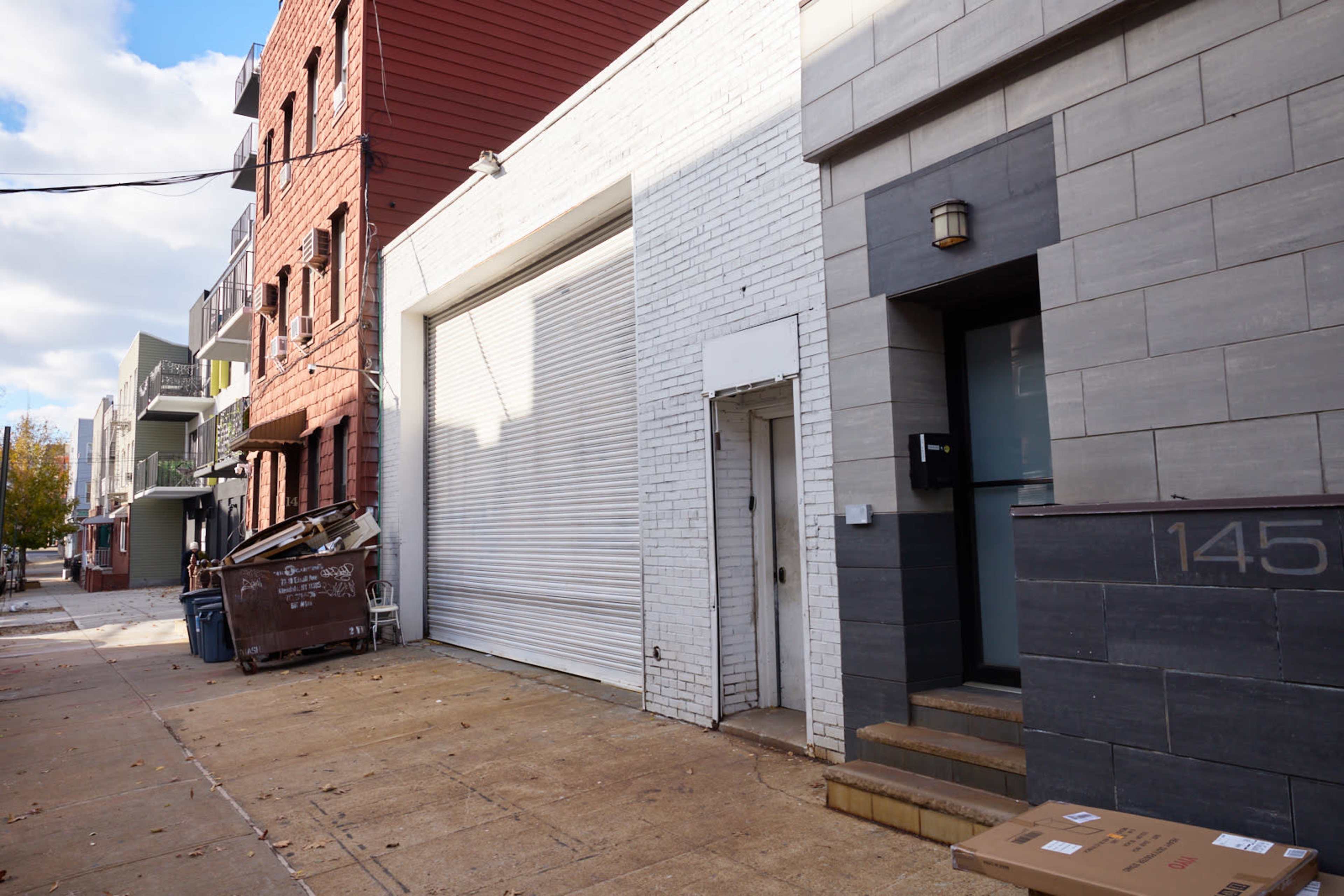 The image shows a street scene featuring a white building with a large roll-up garage door, adjacent to a gray facade building with a front entrance, alongside red-brick and green-sided structures.