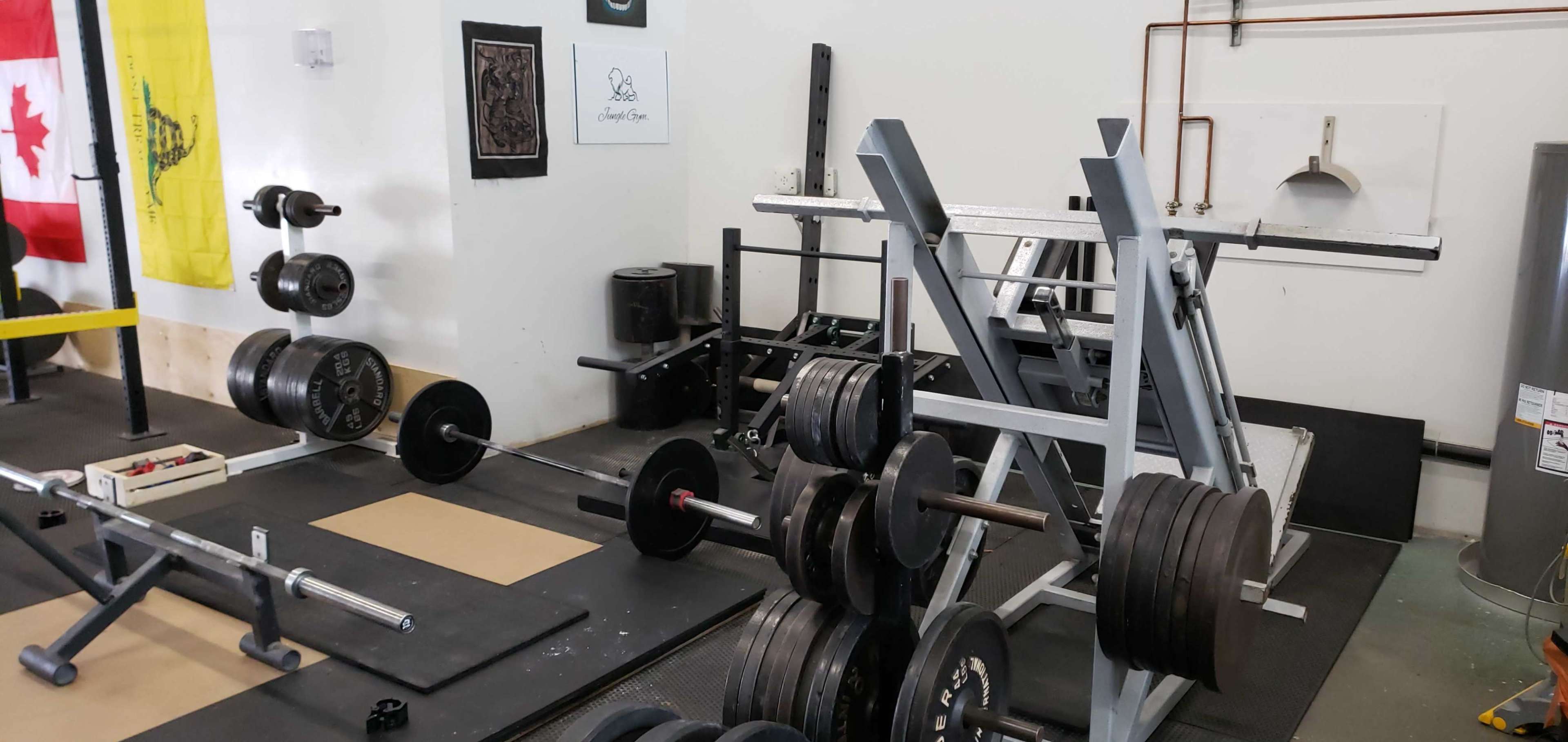 A weightlifting area with various exercise equipment, including a squat rack, free weights, and a barbell set against a wall decorated with flags and artwork.