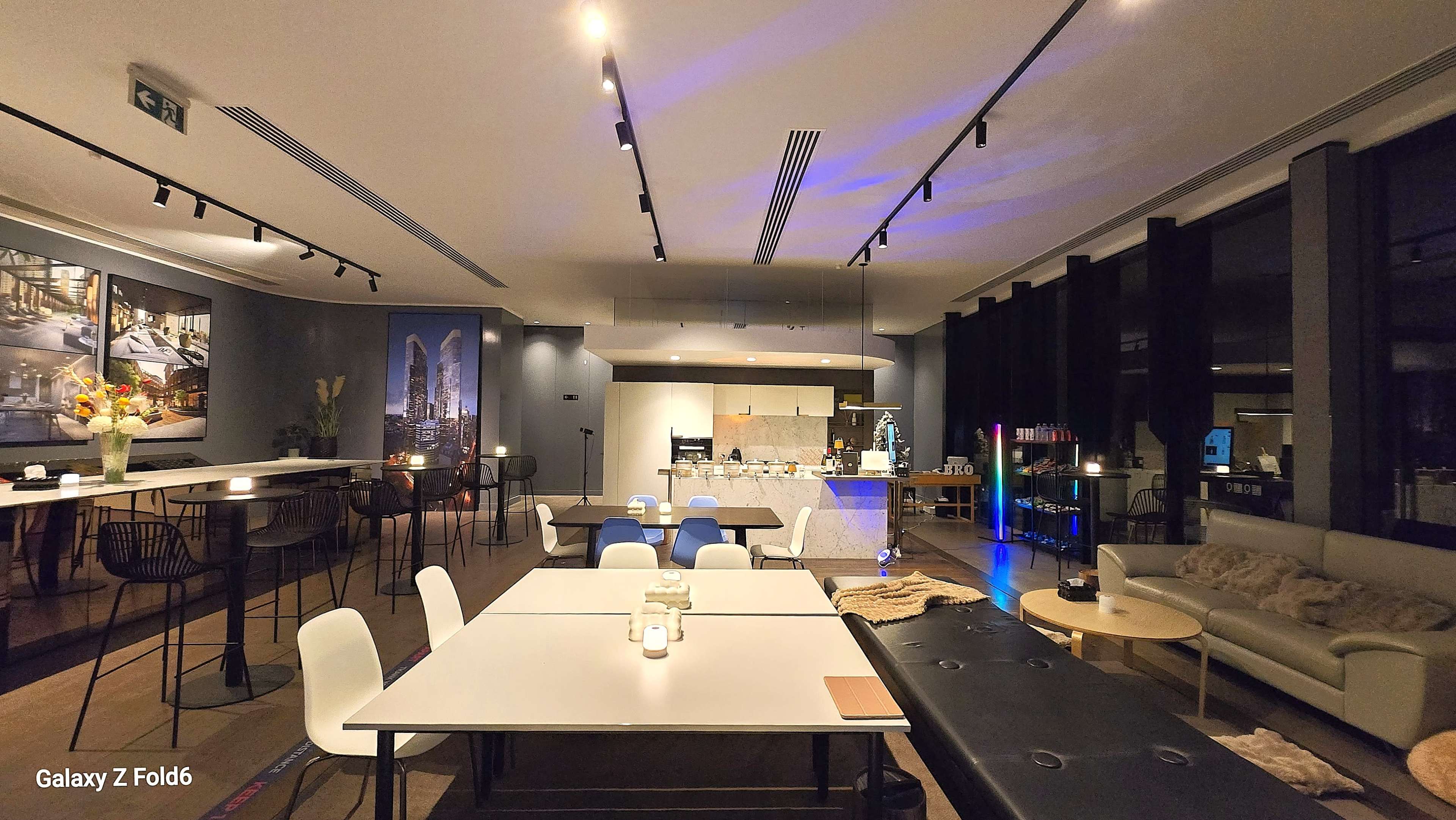 The image shows a modern, well-lit interior space with tables and chairs arranged for dining, featuring a bar area and a kitchen in the background.