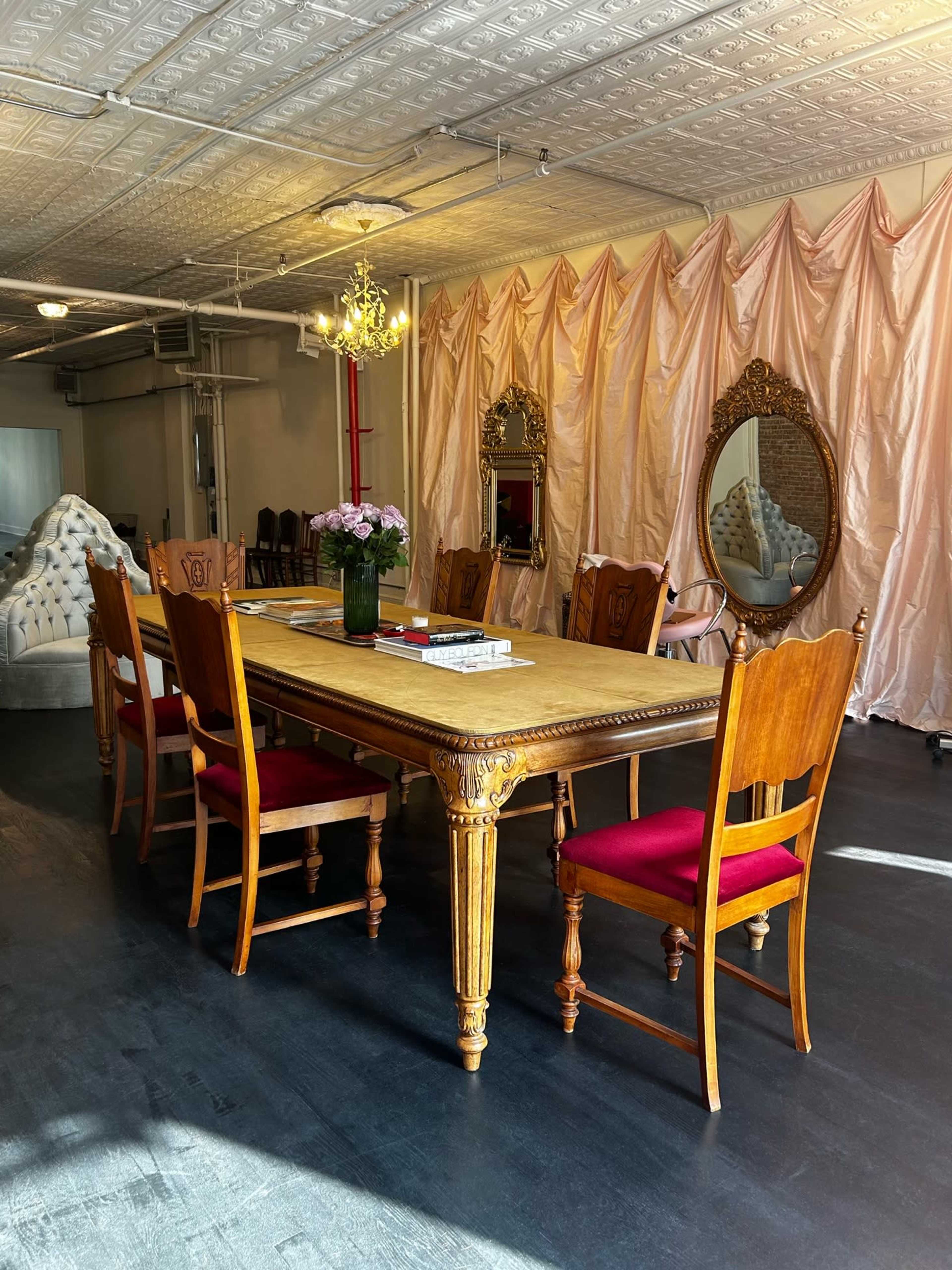 A long wooden dining table is surrounded by eight chairs in a well-lit room with mirrored accents and a fabric drape in the background.