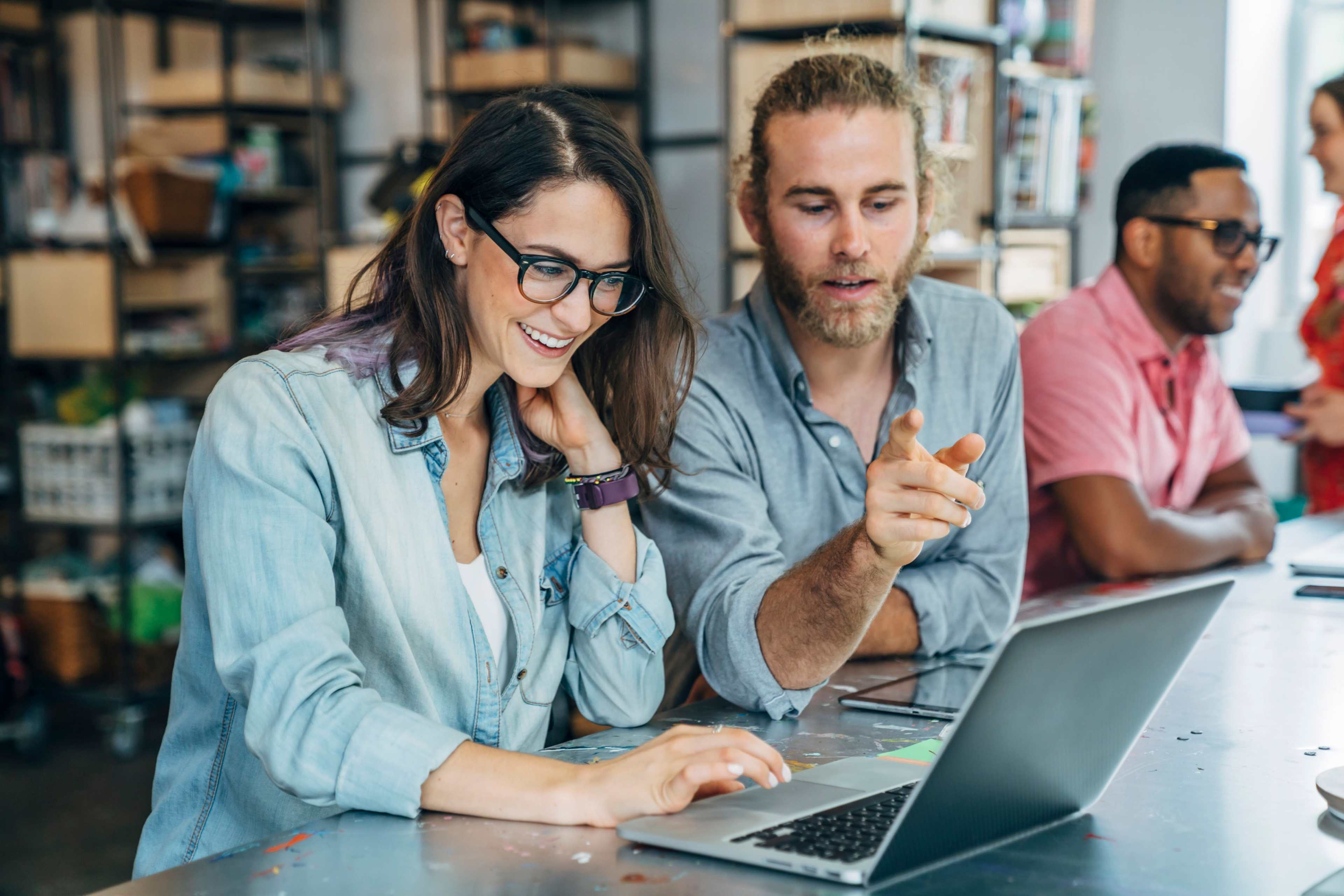 A woman and a man collaborate on a laptop while two other individuals engage in conversation in a modern workspace.