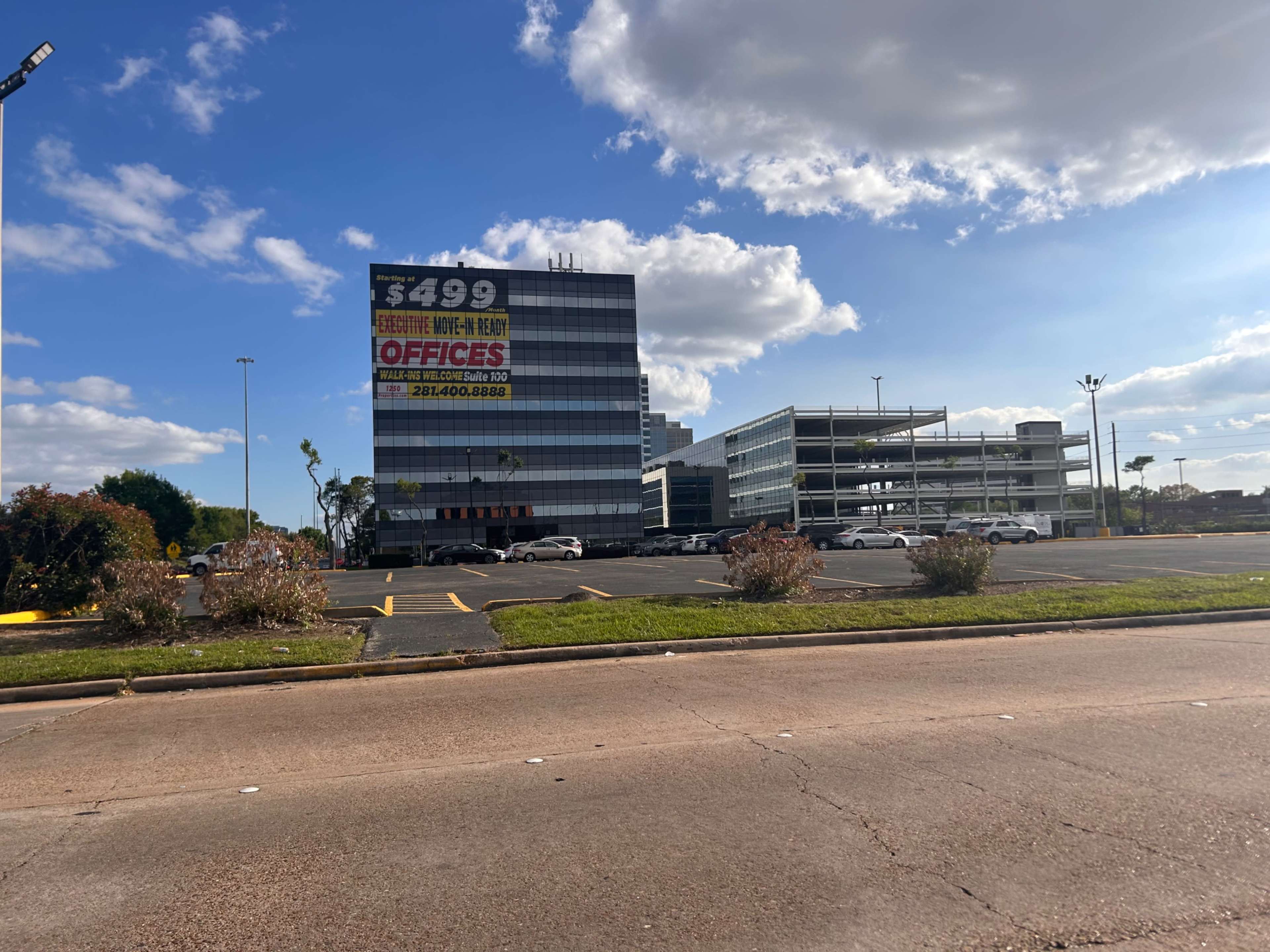 The image shows a large multi-story office building with signage advertising available office spaces for rent, set against a clear blue sky with a few clouds.