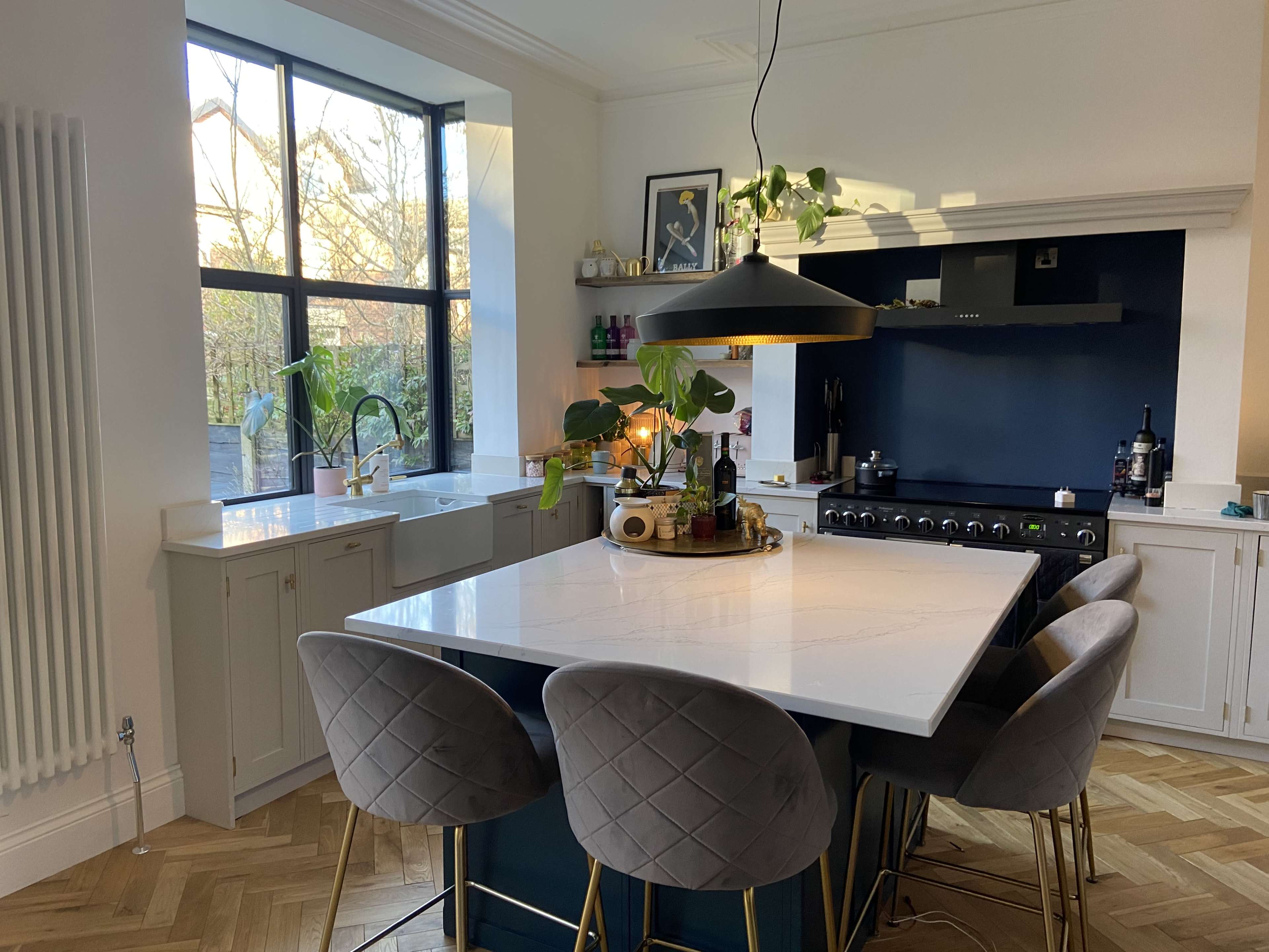 The image shows a modern kitchen featuring a large white island with four velvet bar stools, a dark blue wall cabinet, and large windows that provide natural light.