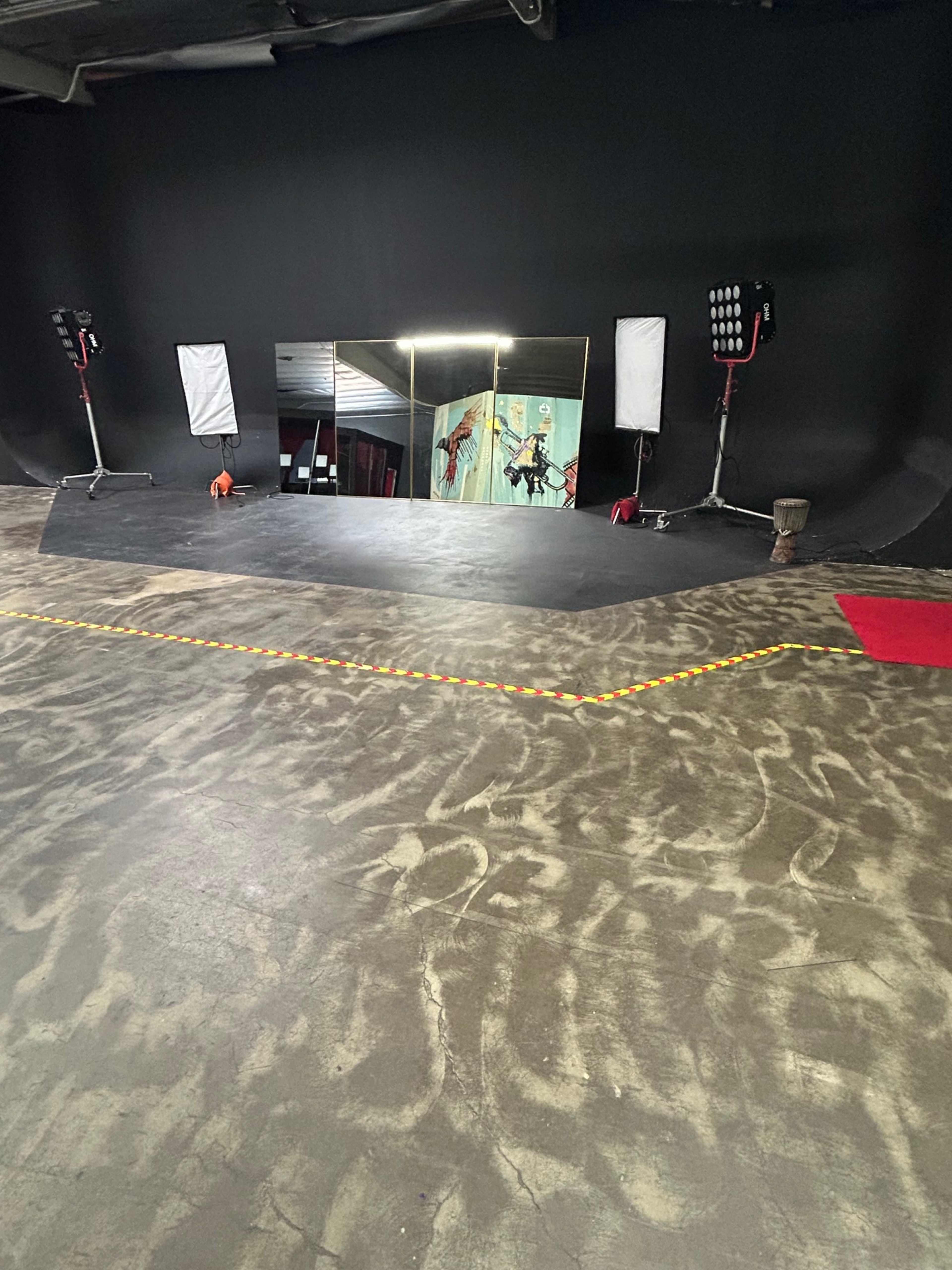 The image shows a stage area with a black backdrop, two lights, and a large mirror, with red and yellow tape marking a border on the floor.