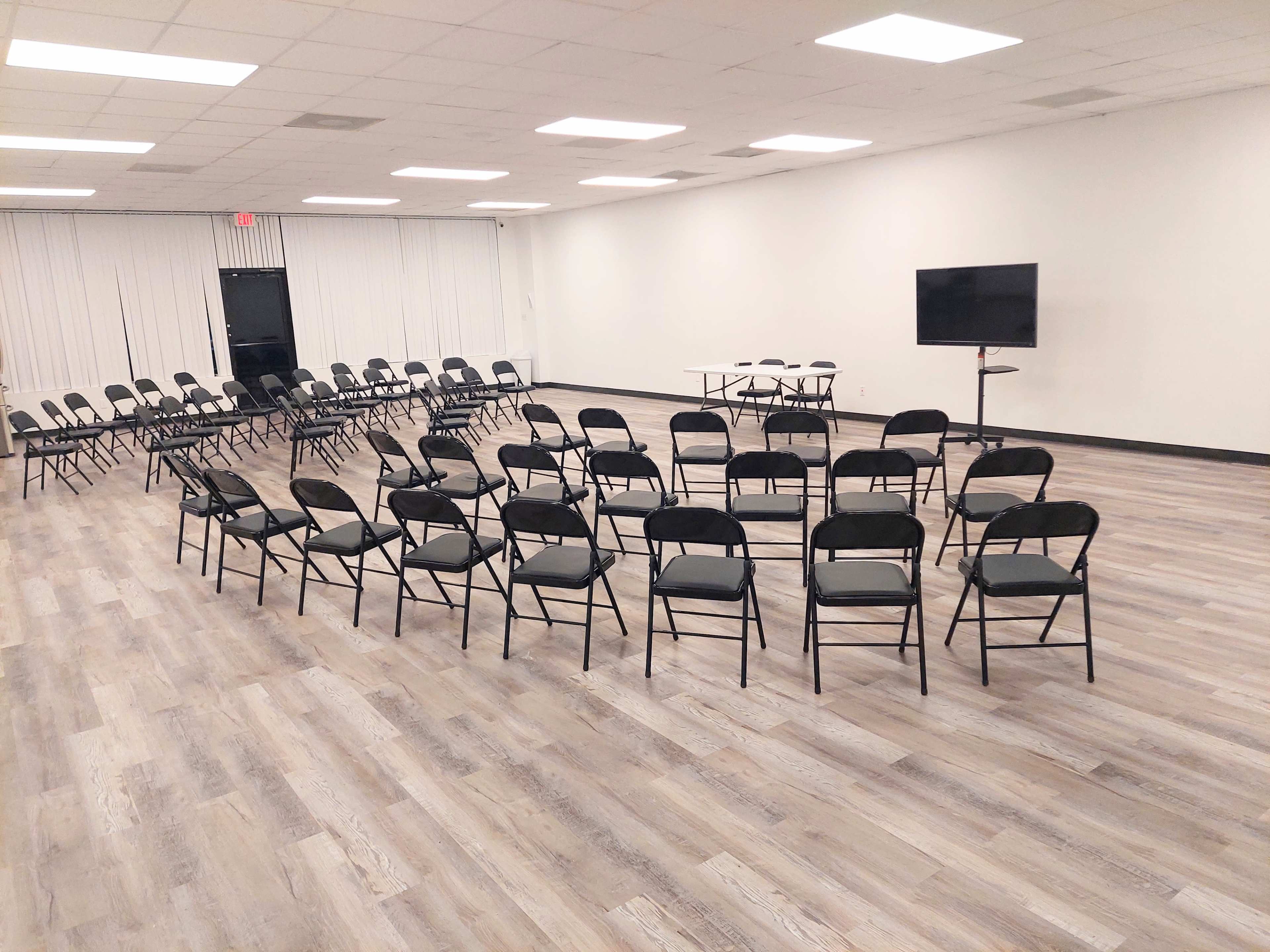 A spacious indoor setting with rows of black folding chairs arranged in a circle, surrounding a central area with a table and a television.