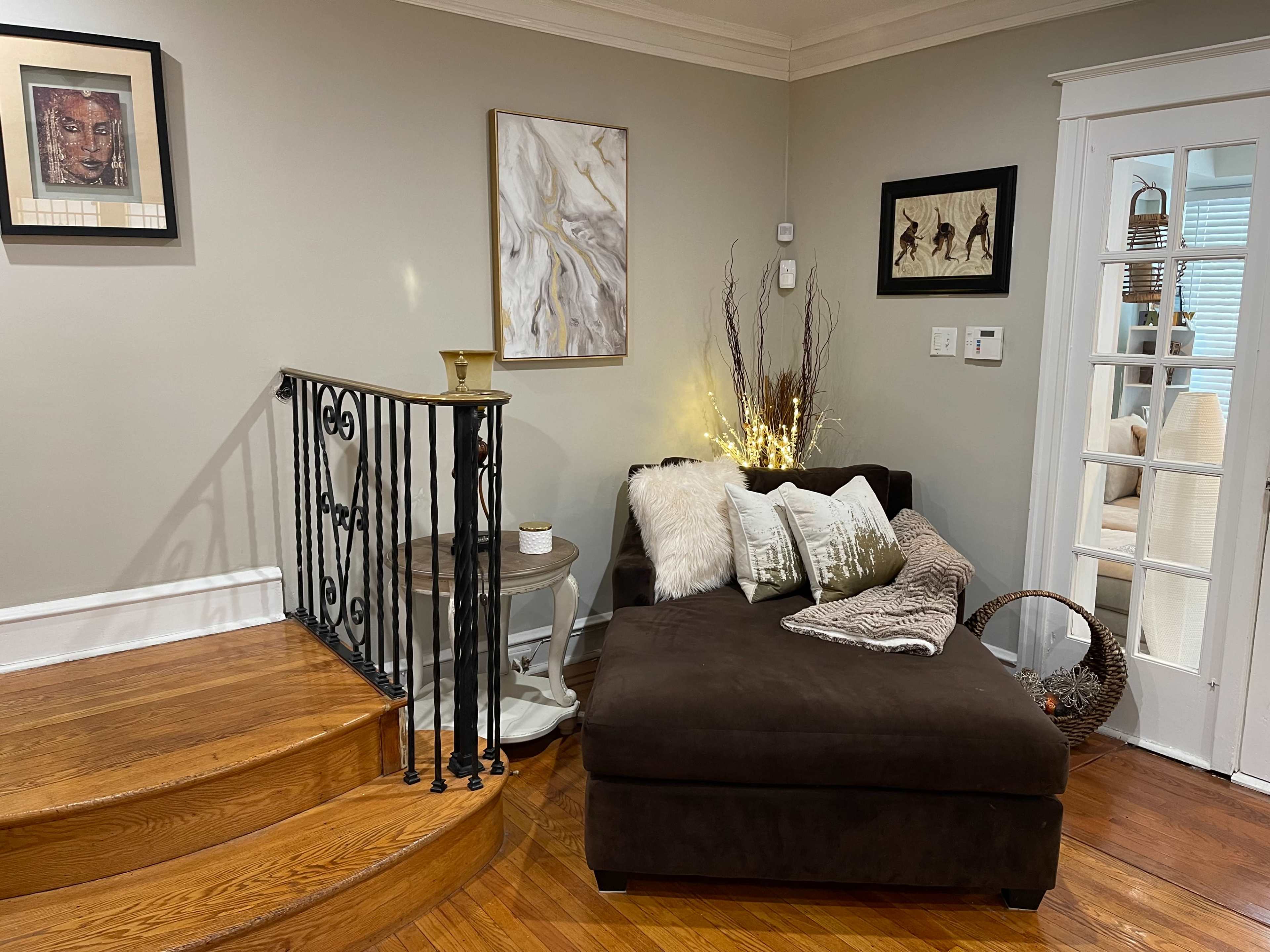 A cozy living space featuring a dark chaise lounge with decorative pillows, a small side table, and a staircase with a wrought iron railing against a light-colored wall.