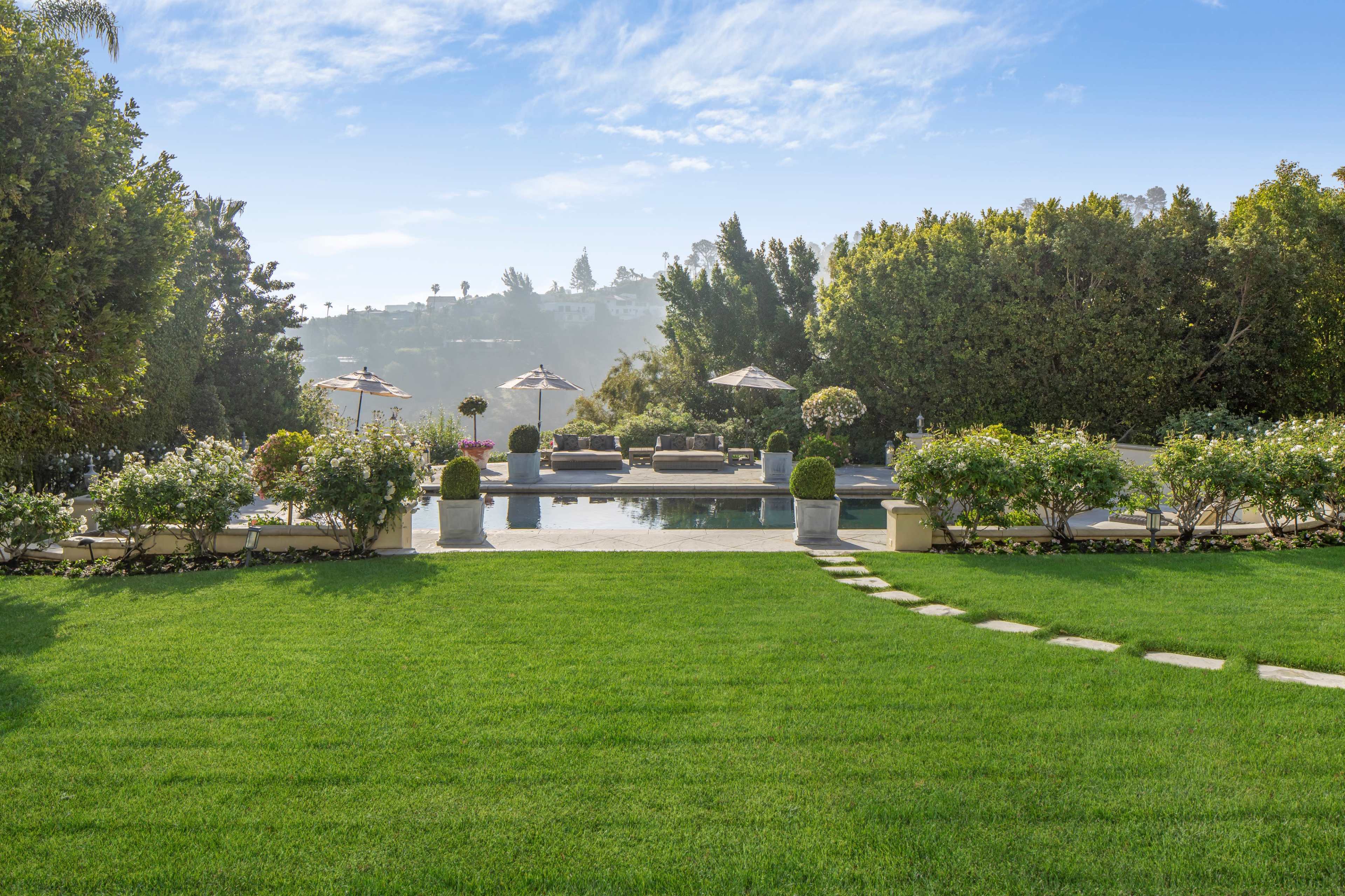 The image shows a spacious backyard with a well-manicured lawn, a swimming pool, and seating areas shaded by umbrellas.
