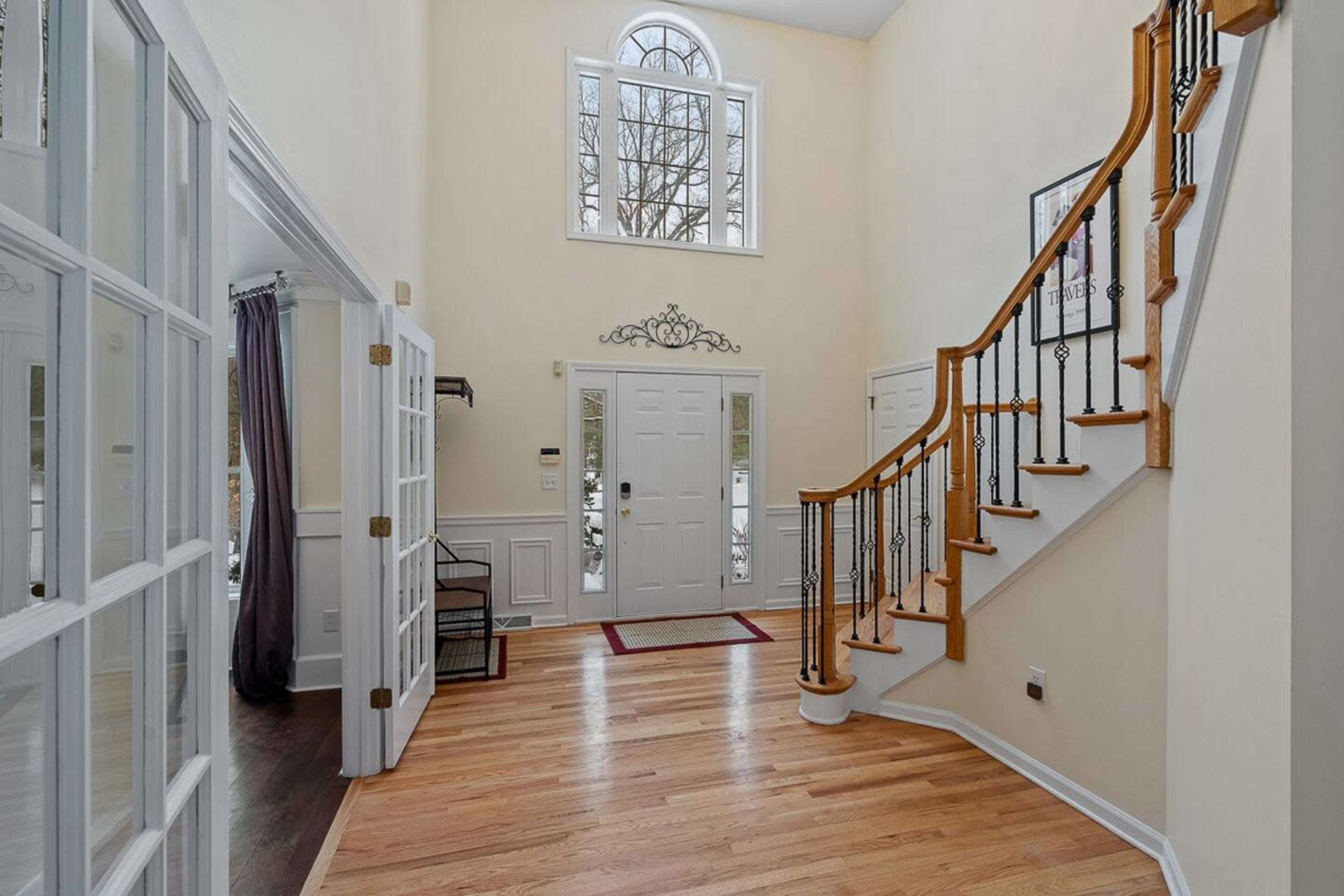 A spacious foyer with a staircase, large windows, and double doors leading outside, featuring hardwood flooring and wall paneling.