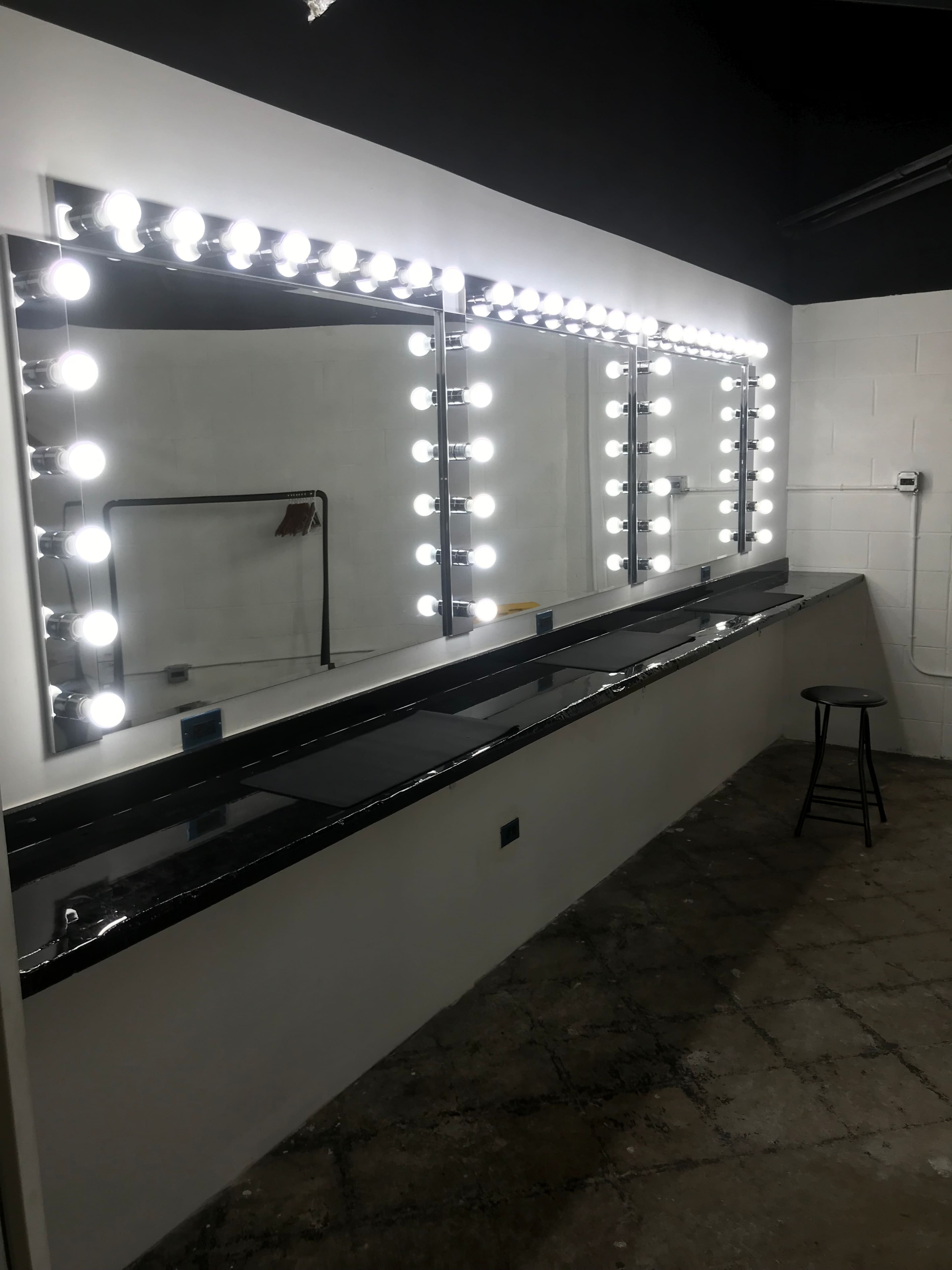A spacious vanity area with bright, circular lights surrounding three large mirrors above a black countertop and a single stool.
