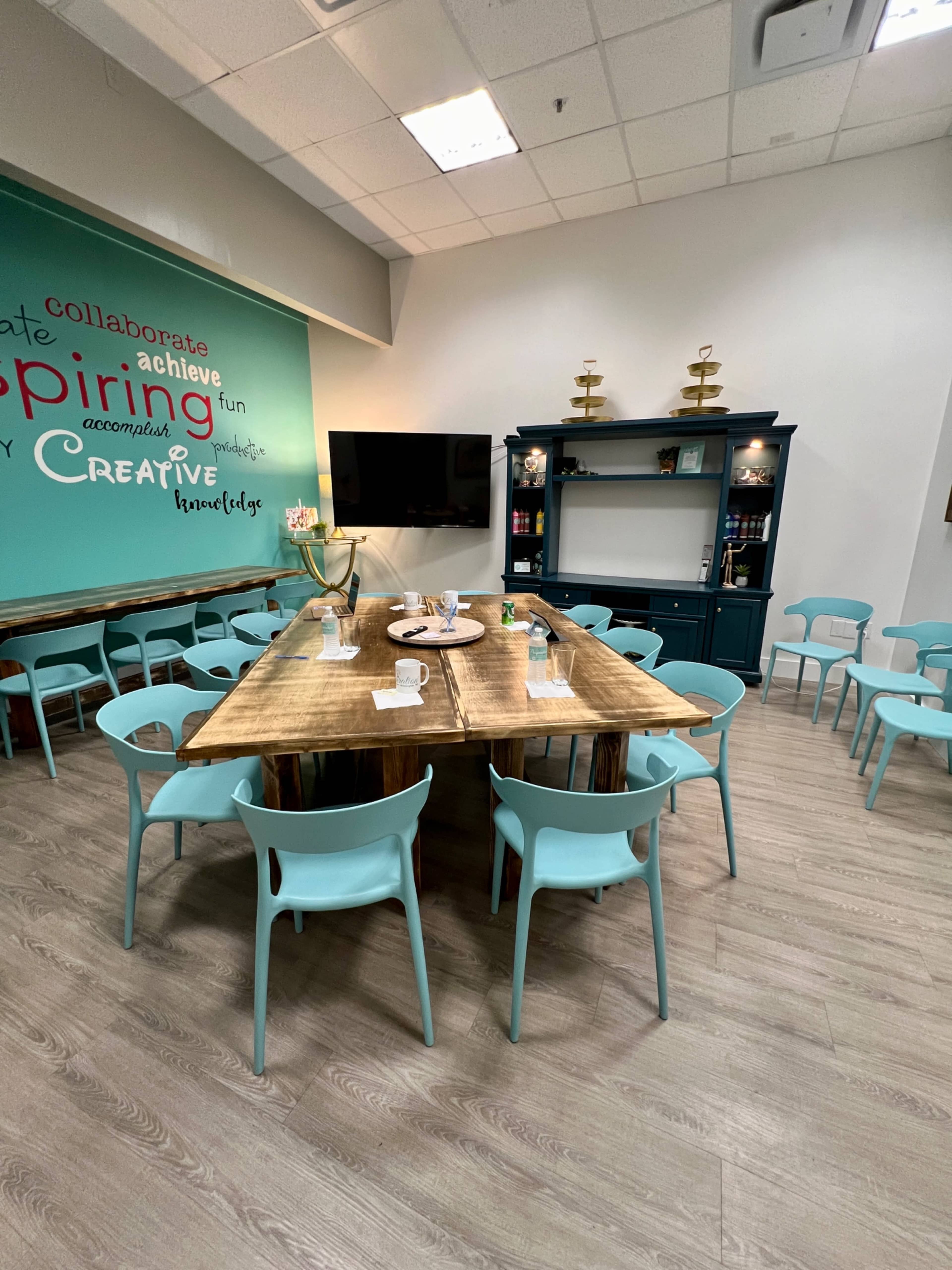 A spacious room with a large wooden table surrounded by light blue chairs, a TV mounted on the wall, and a teal cabinet displaying decorative items.