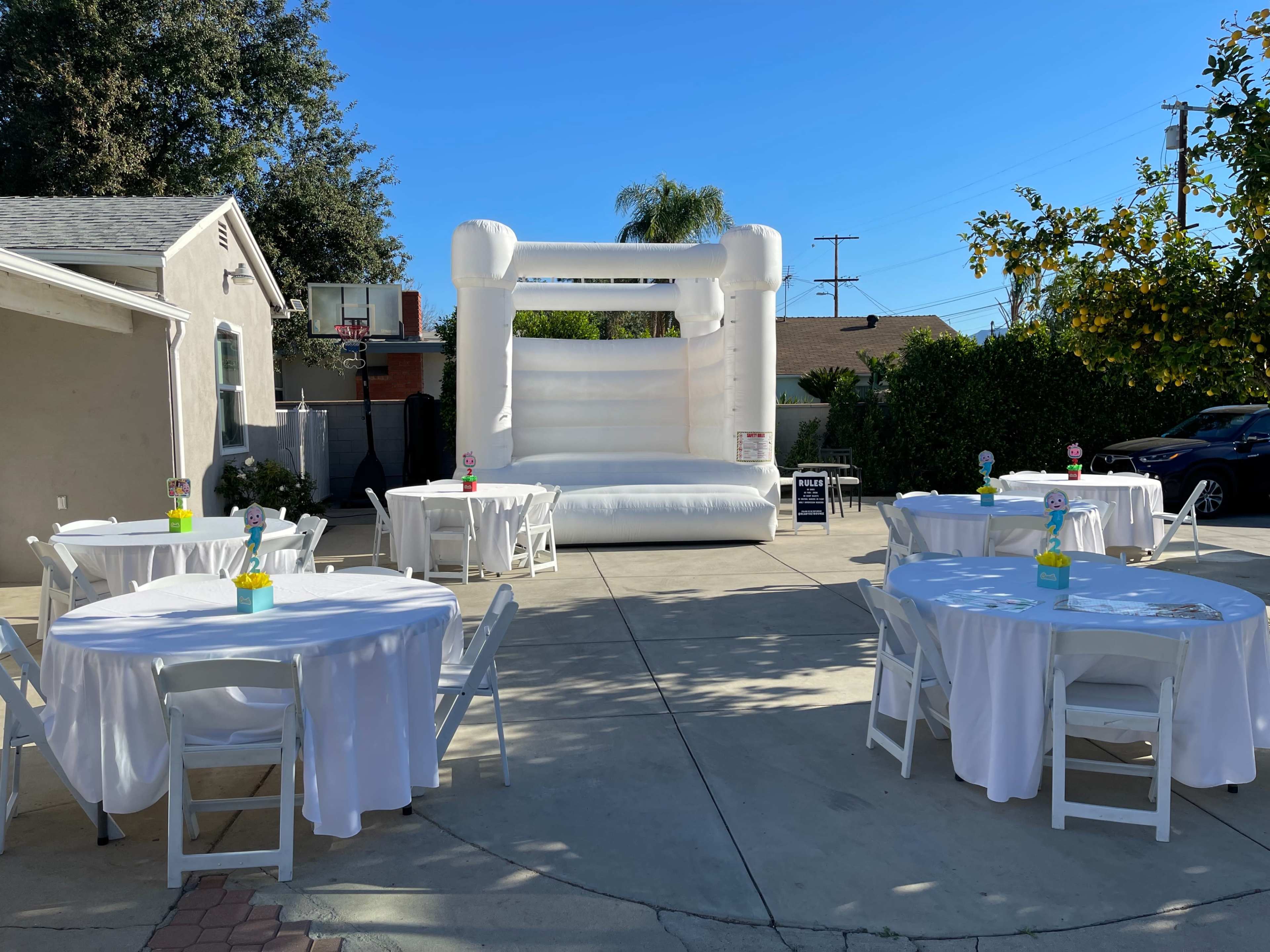 A backyard setting features a white bouncy castle and several round tables covered with white tablecloths.