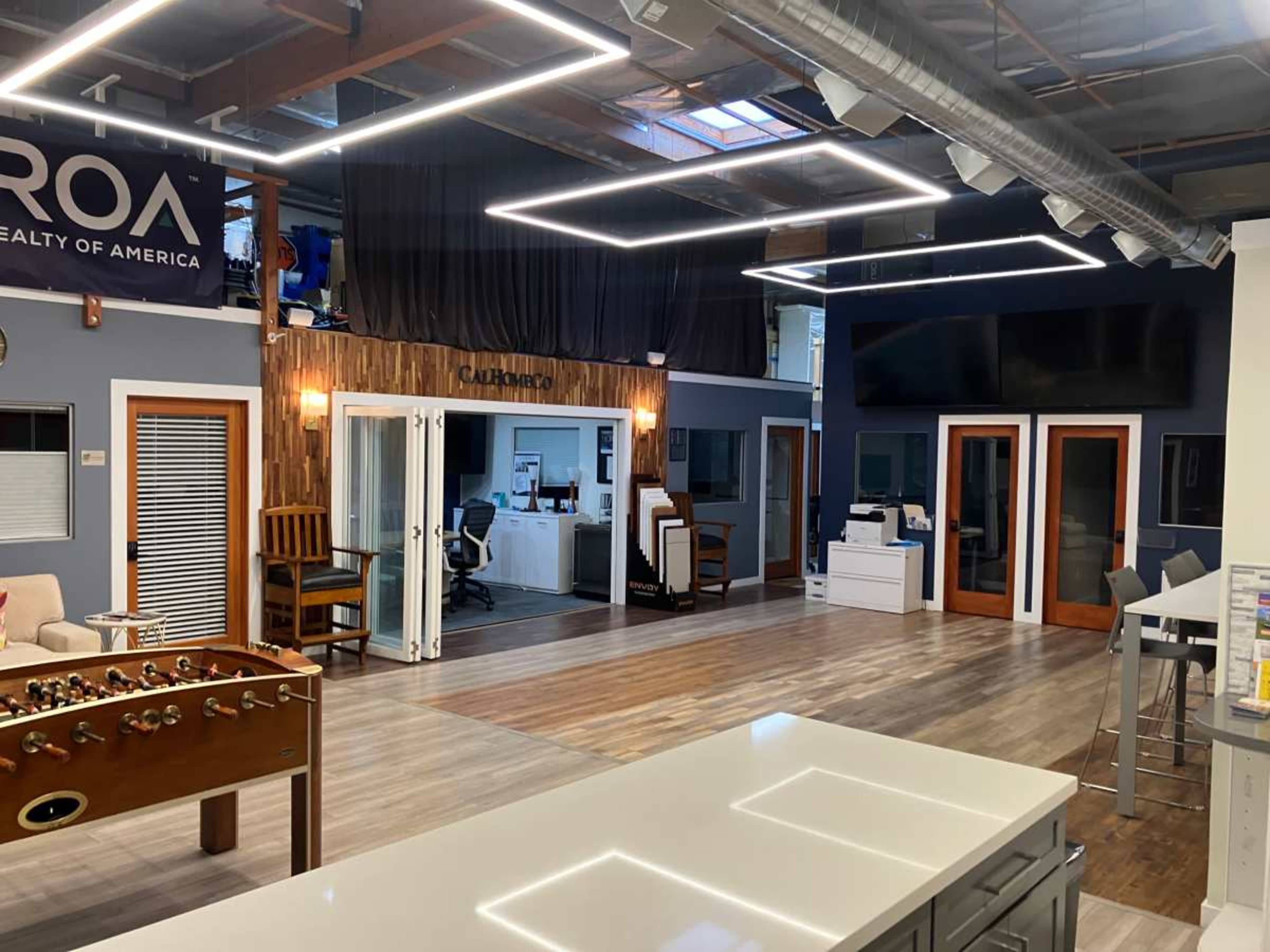 The image shows a modern office space with wooden flooring, features a foosball table, and several workstations, including a reception area and private offices.