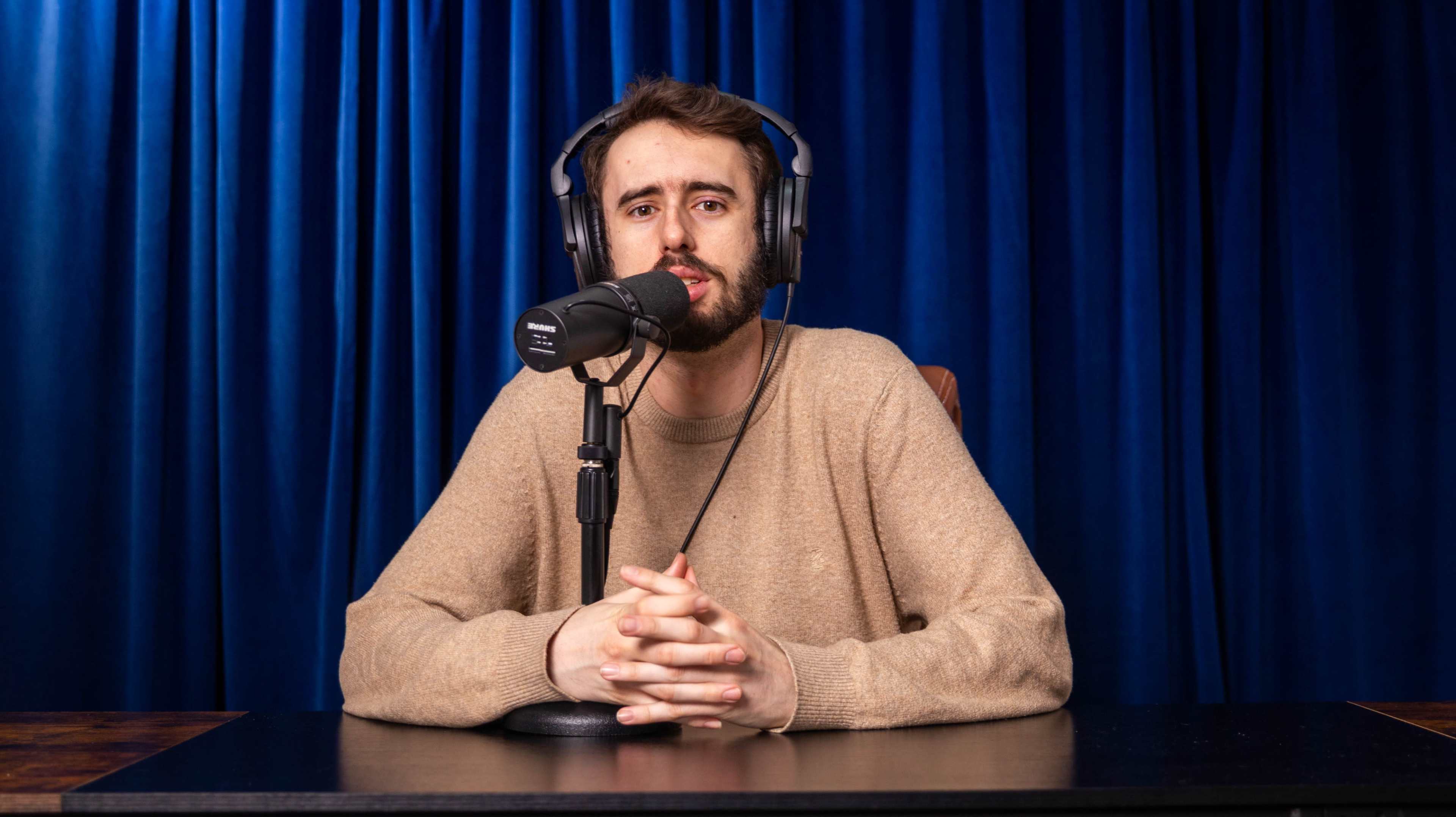 A man wearing headphones sits at a desk with a microphone in front of him, against a blue curtain backdrop.