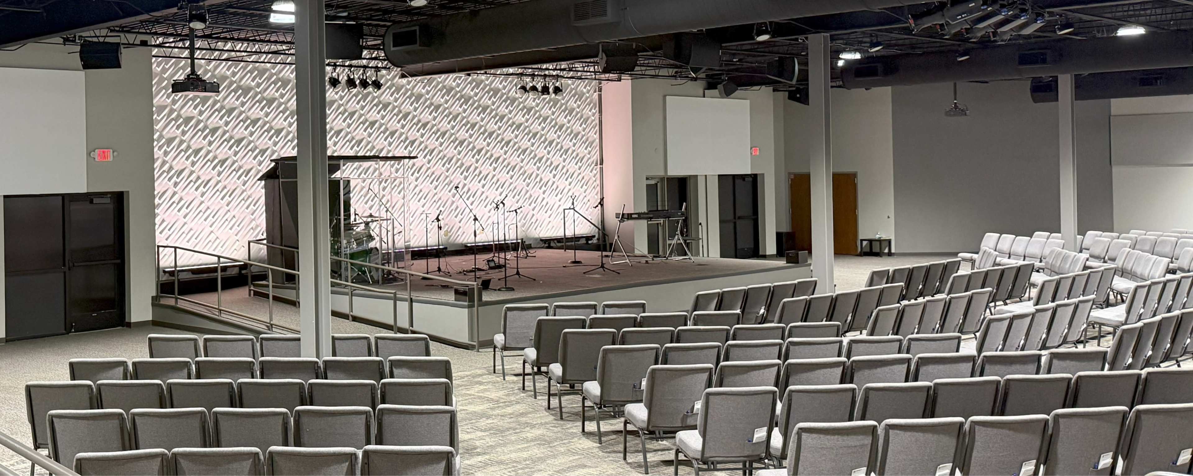 The image shows a spacious auditorium with rows of gray chairs facing a stage equipped with musical instruments and a backdrop displaying a pattern.