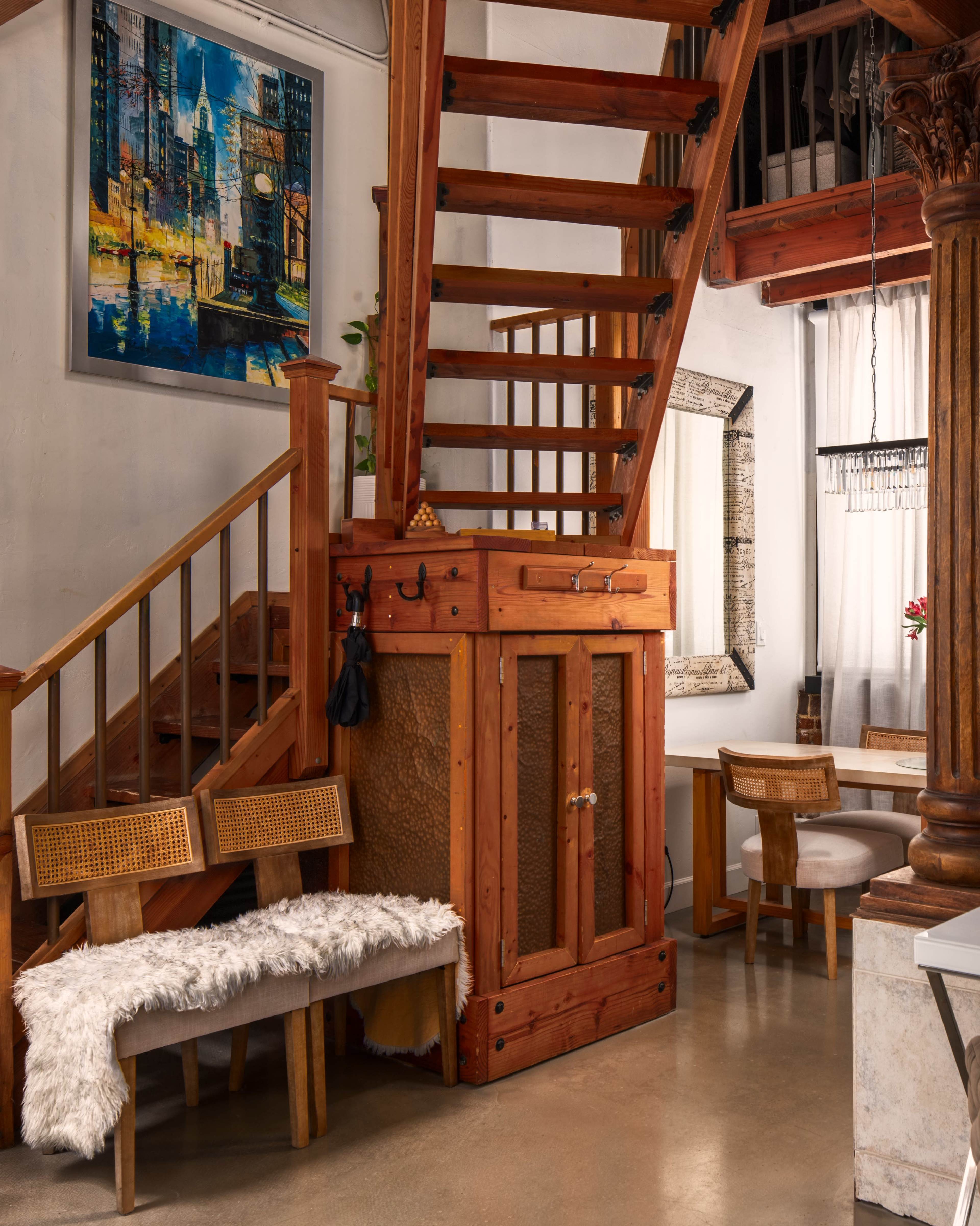 The image features a wooden staircase leading to an upper level in a spacious interior, with a small seating area and a decorative cabinet beneath the stairs.