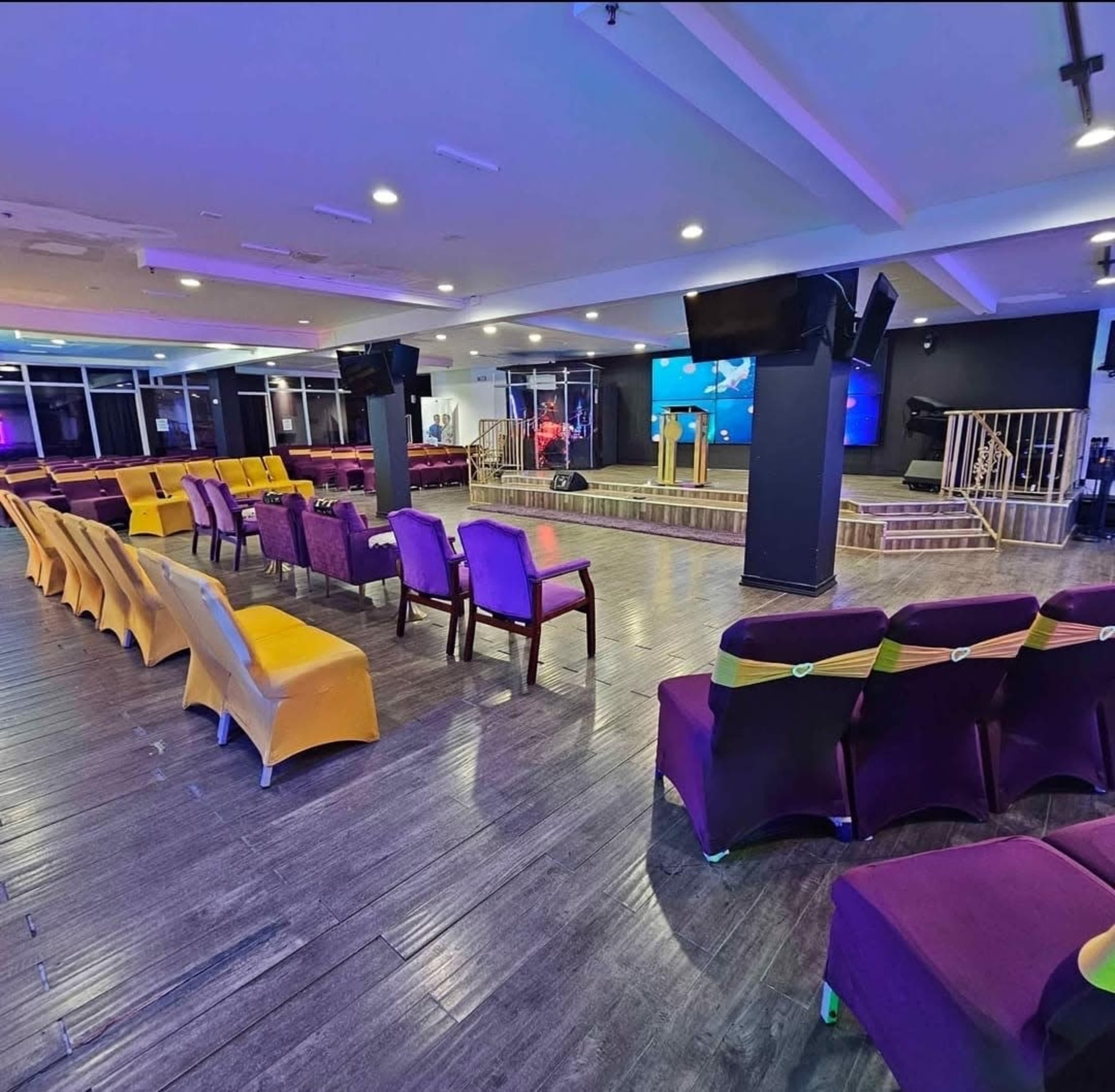 The image shows a modern event space with arranged seating in purple and yellow colors facing a stage with a podium and screens.
