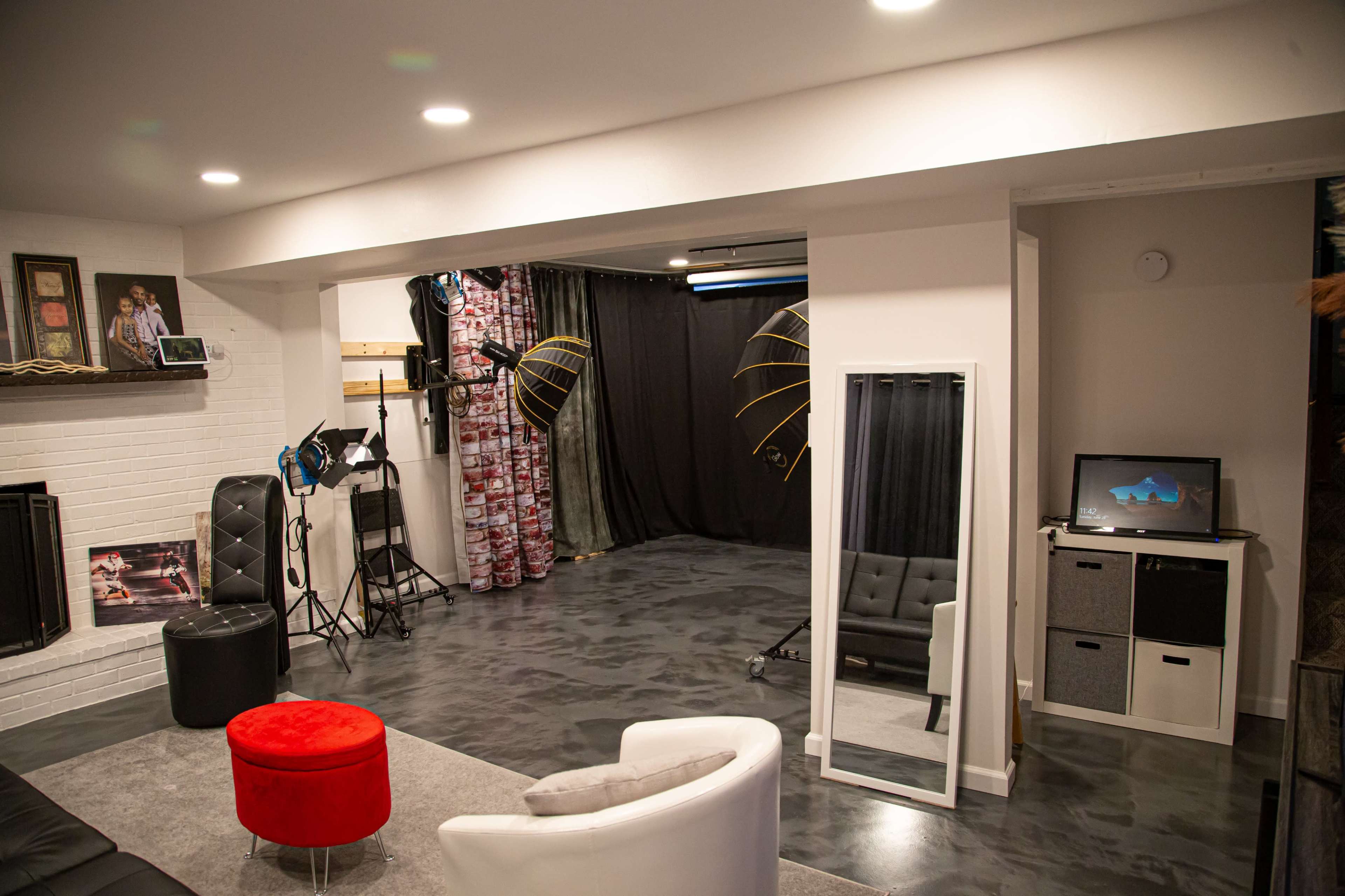 A modern studio space featuring a seating area with a red ottoman, a large mirror, lighting equipment, and a backdrop against a textured floor.