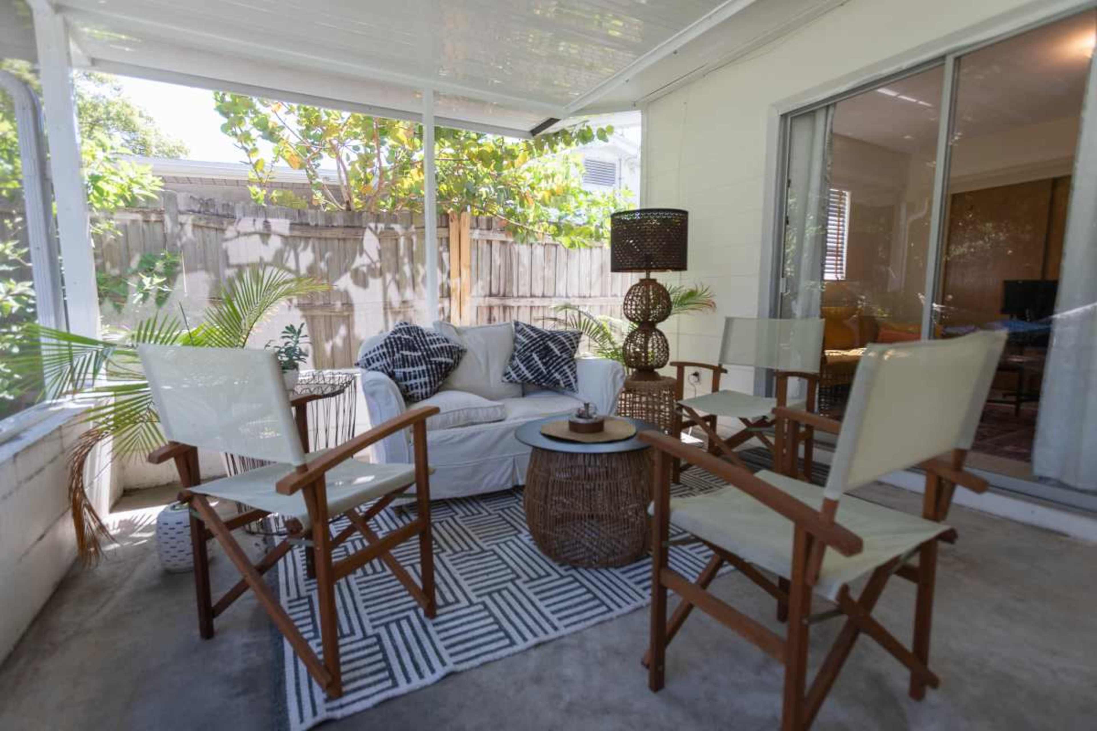 A shaded patio area features a white sofa, four wooden chairs, a round table, and decorative plants.