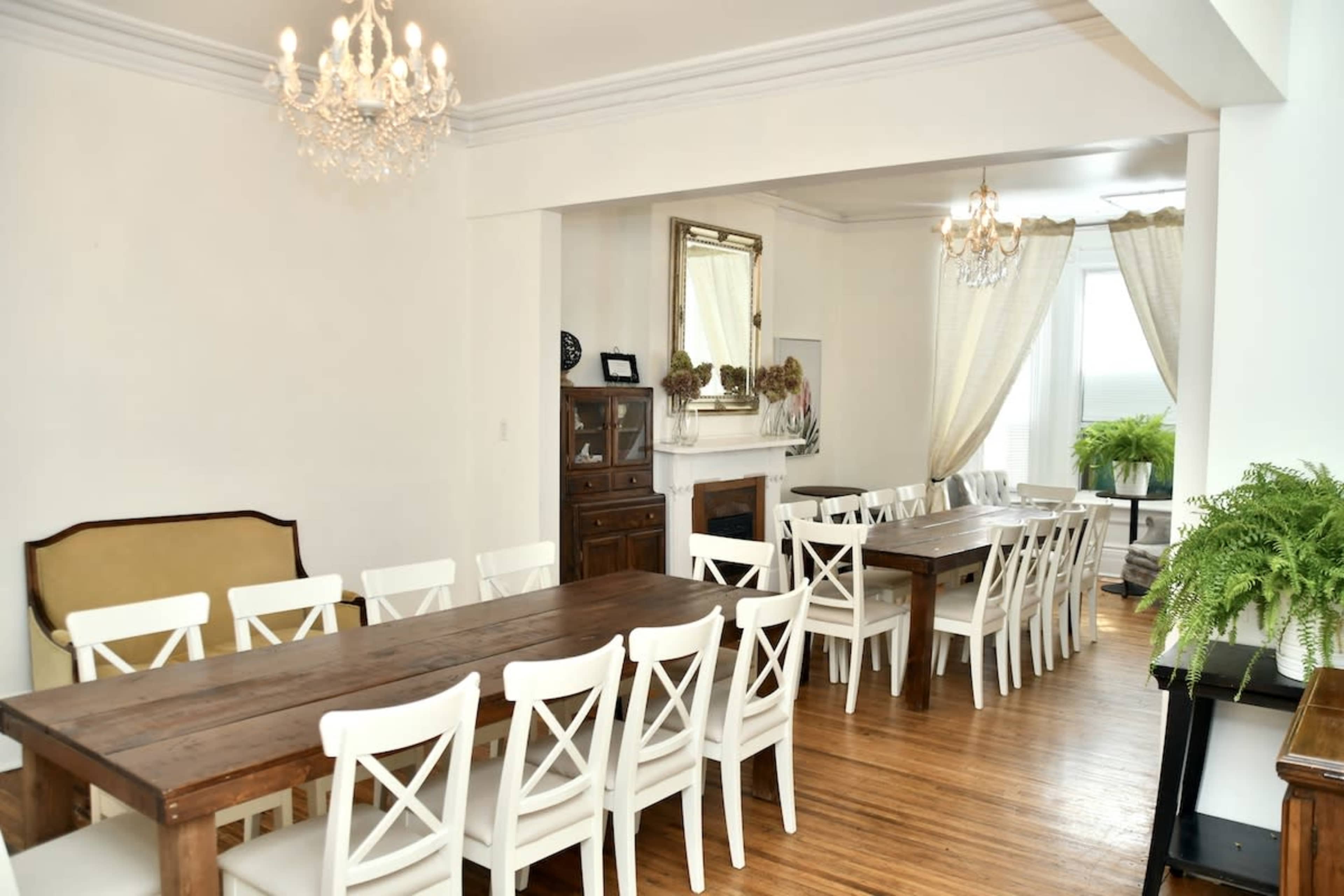 A bright dining area features two long wooden tables with white chairs, chandeliers overhead, and large windows draped with sheer curtains.