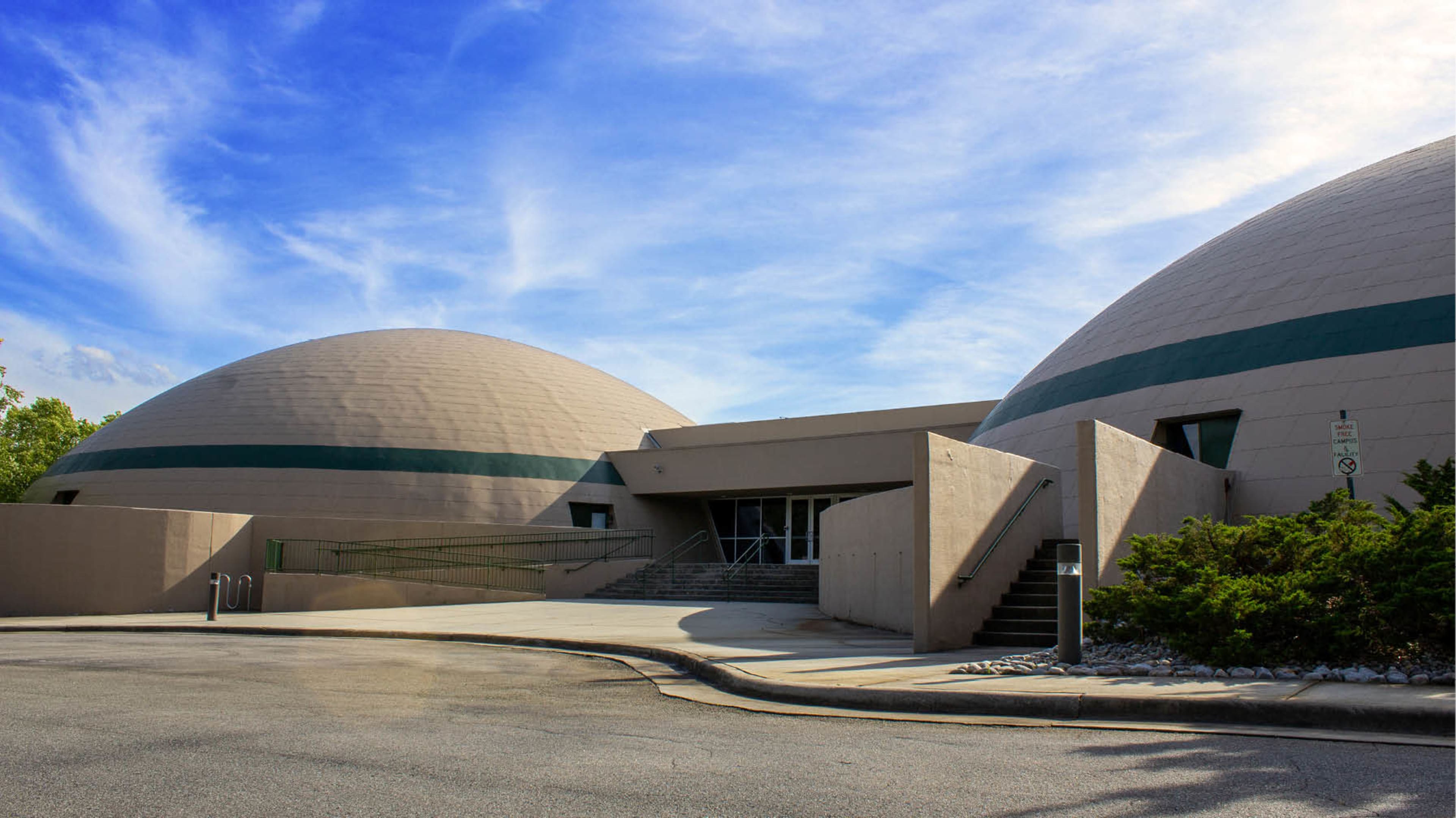 The image shows a modern building featuring two large dome structures and a concrete entrance with steps leading up to it, under a blue sky with clouds.