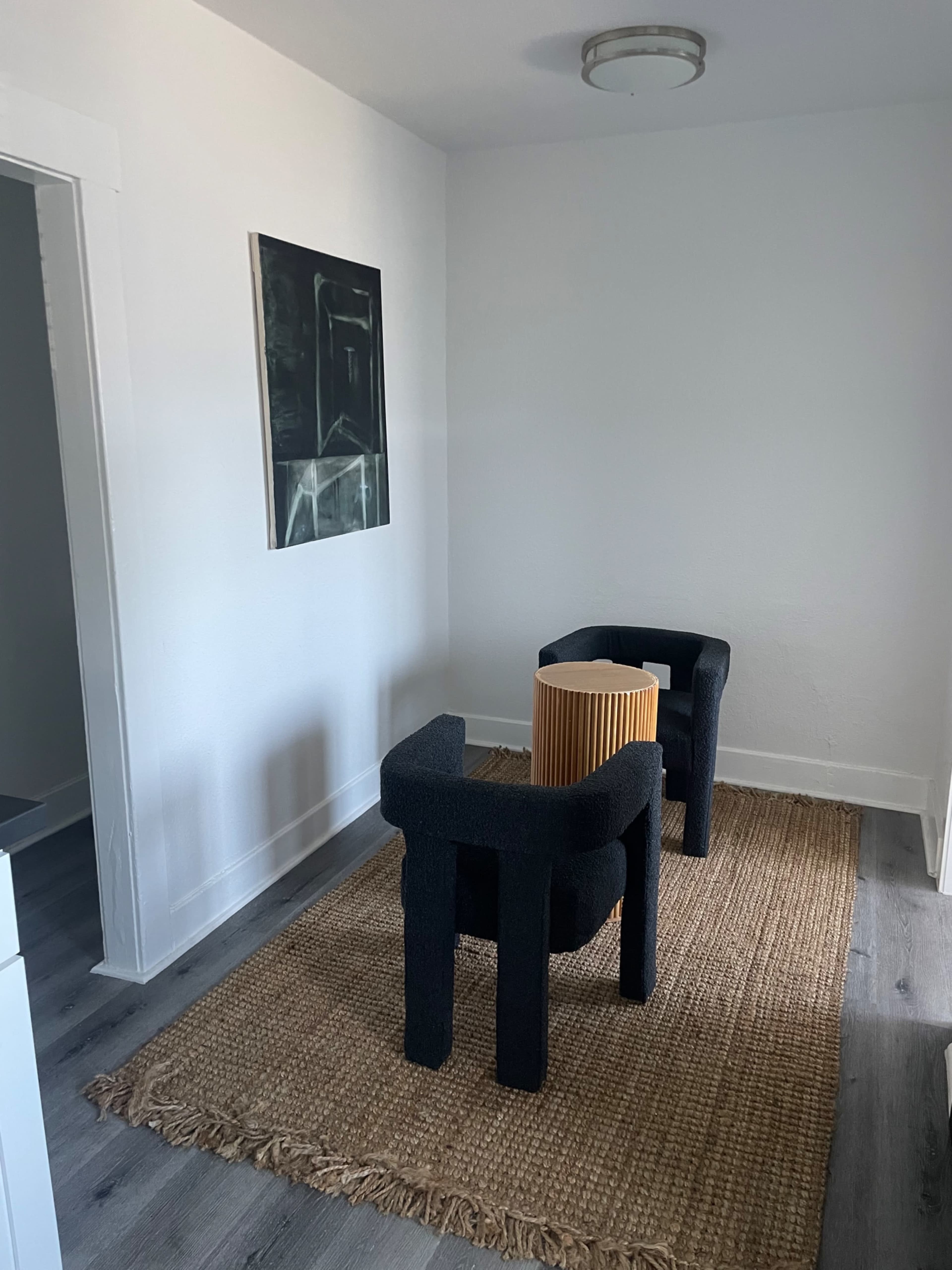 A small, minimally furnished room features two black chairs flanking a cylindrical wooden table on a textured rug.