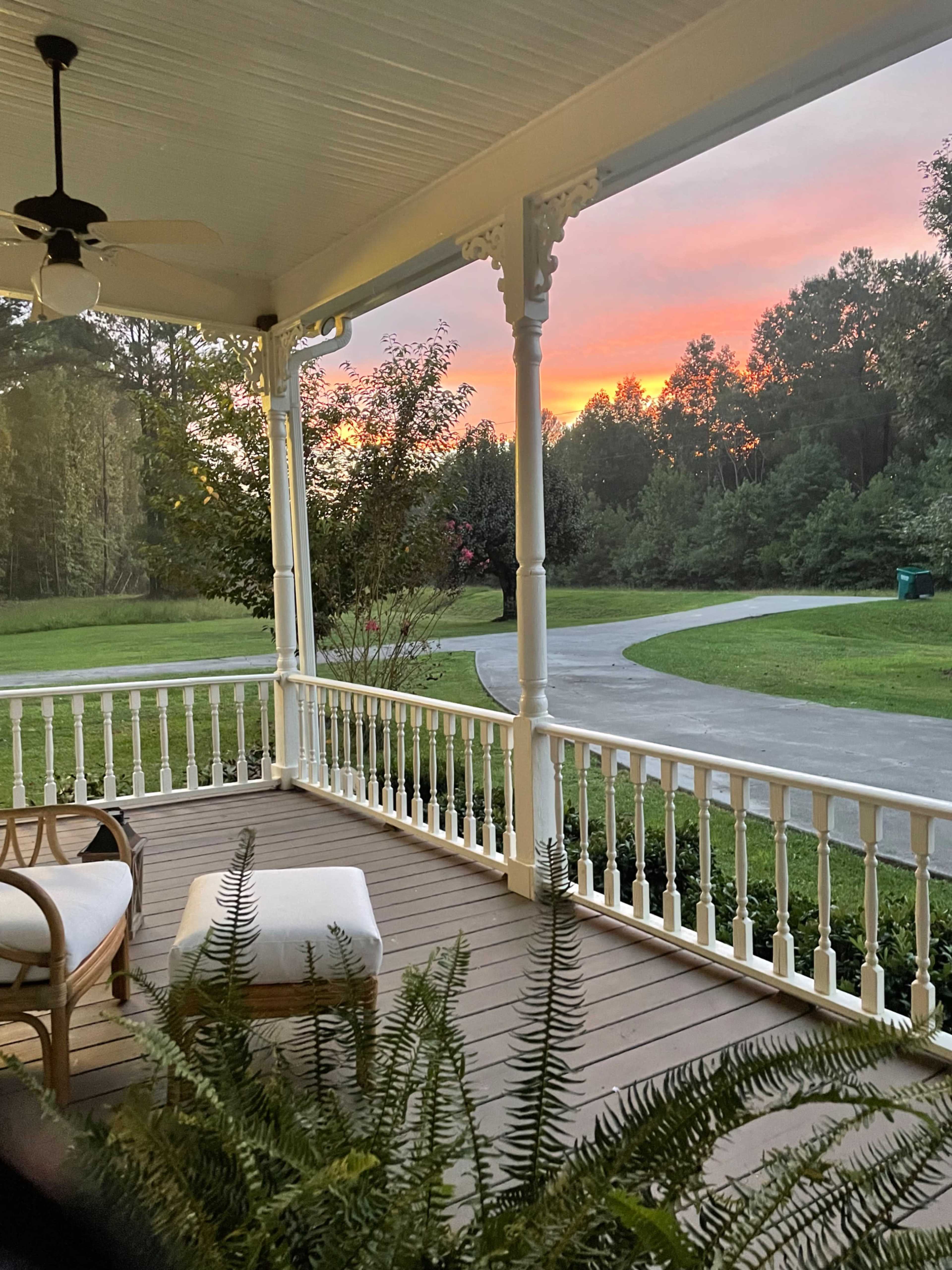 A porch with a rattan chair and white cushion, overlooking a curving driveway and trees at sunset.