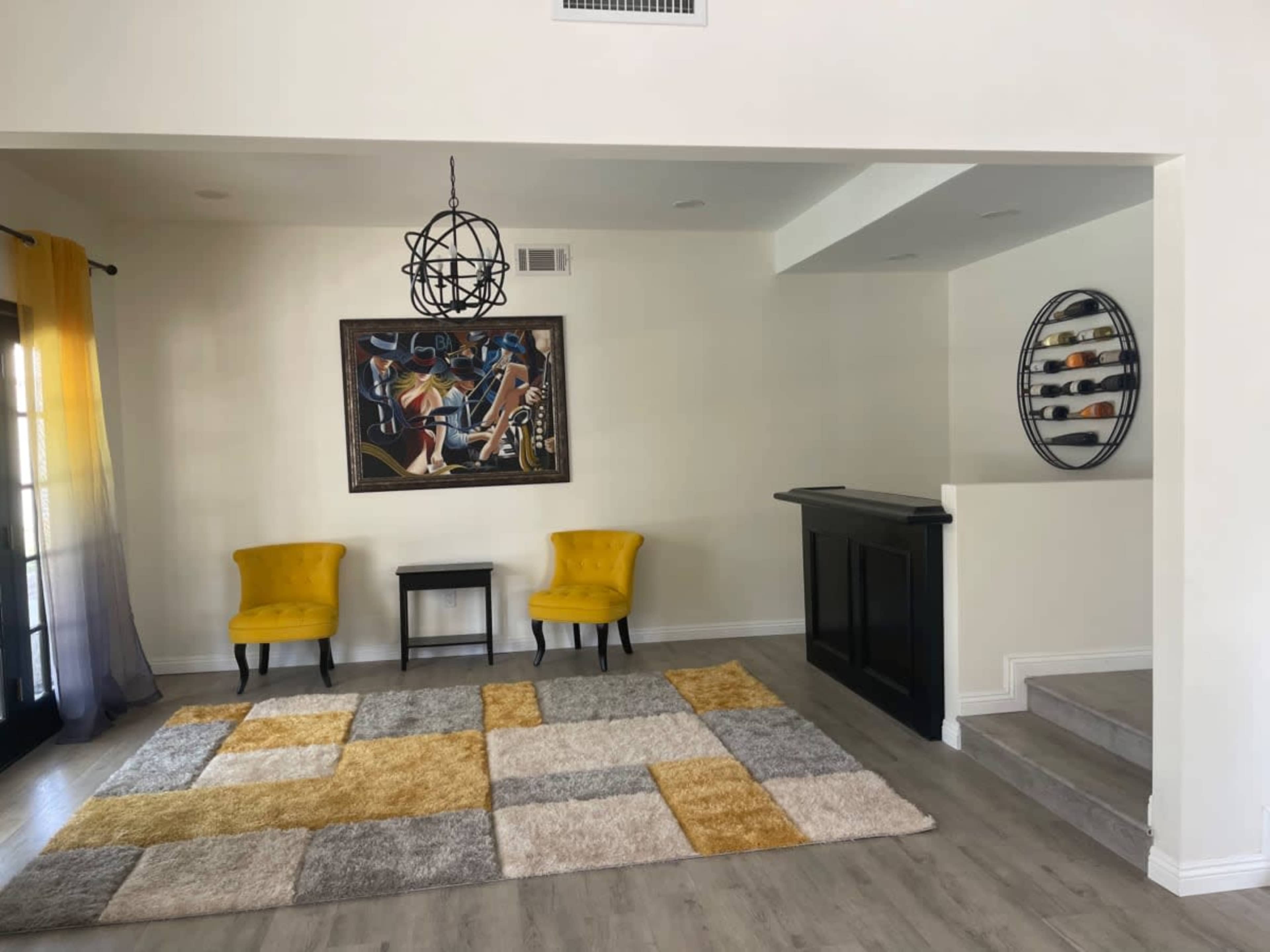 A bright living space featuring two yellow chairs, a small black table, a colorful patchwork rug, and a wall-mounted wine rack.