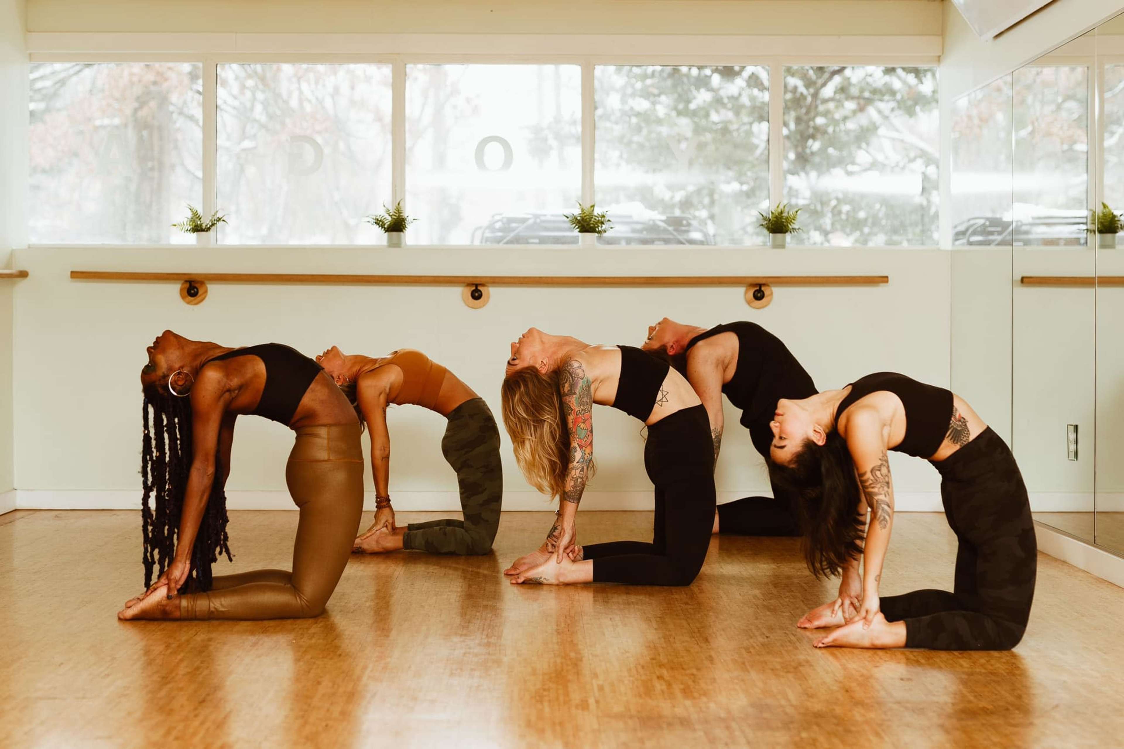 A group of five individuals performs a yoga pose in a brightly lit studio with large windows.