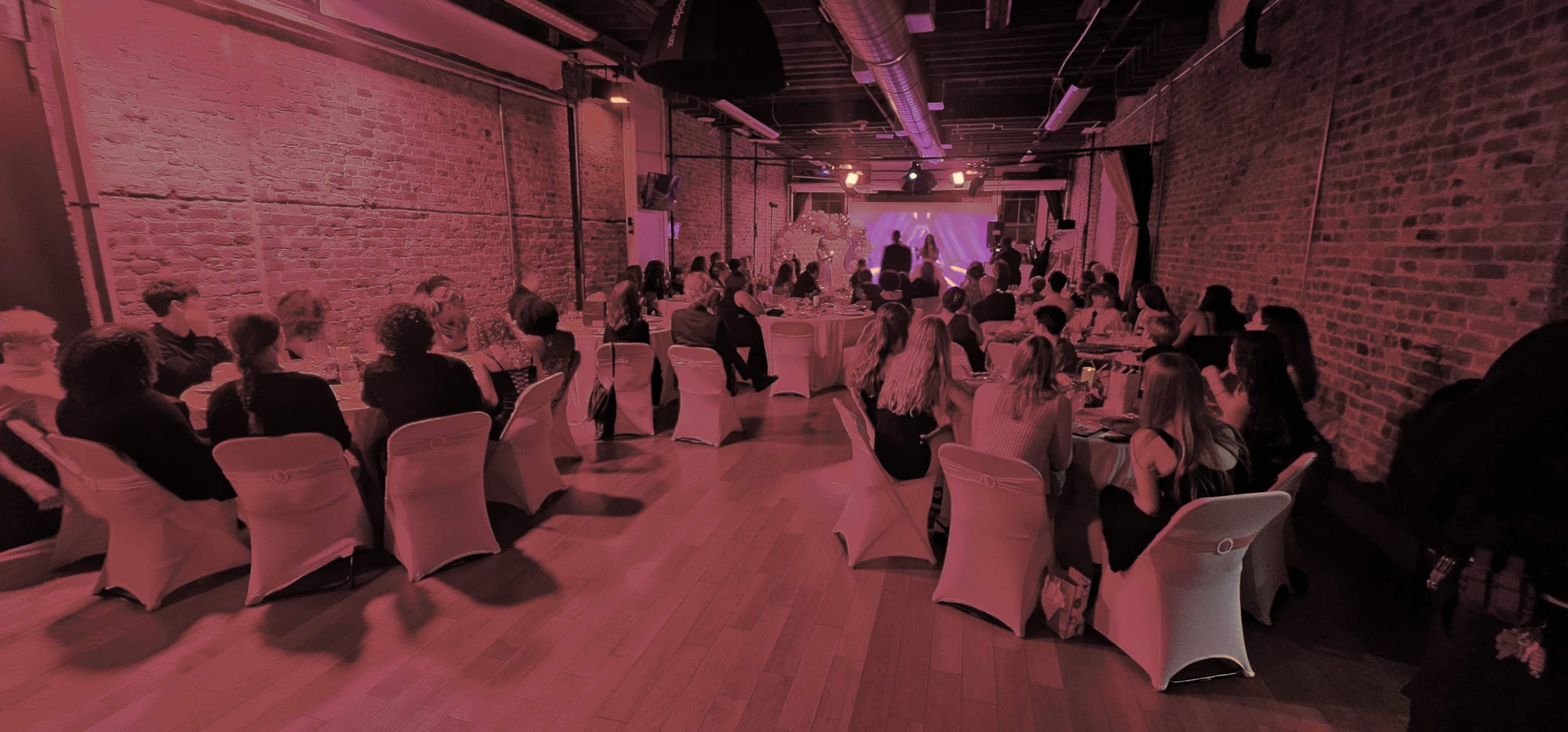 The image shows a gathering in a spacious venue featuring tables with guests seated, facing a stage with performances or presentations.
