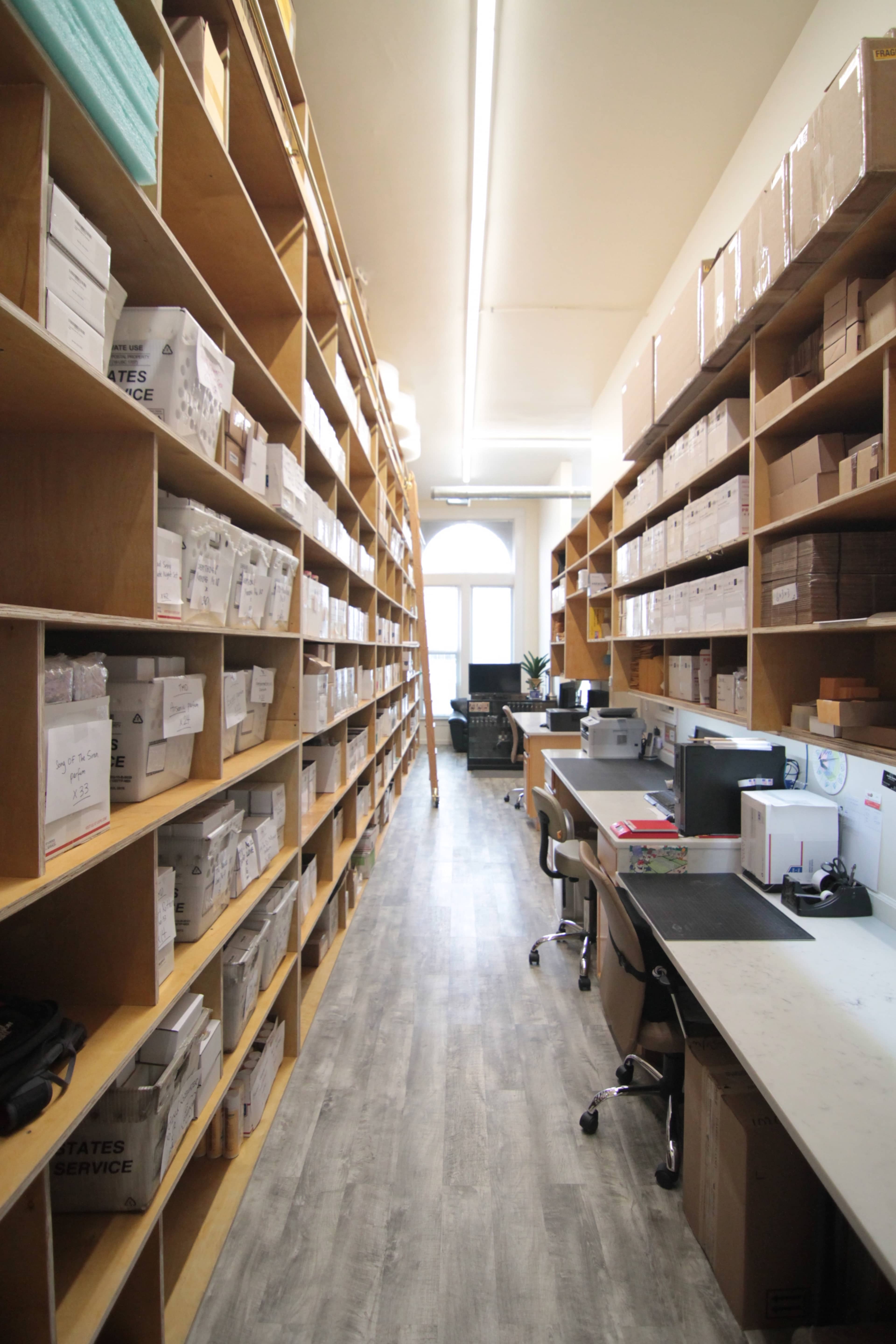 A narrow corridor lined with tall wooden shelves filled with labeled boxes leads to a workspace with desks and computers at the end.