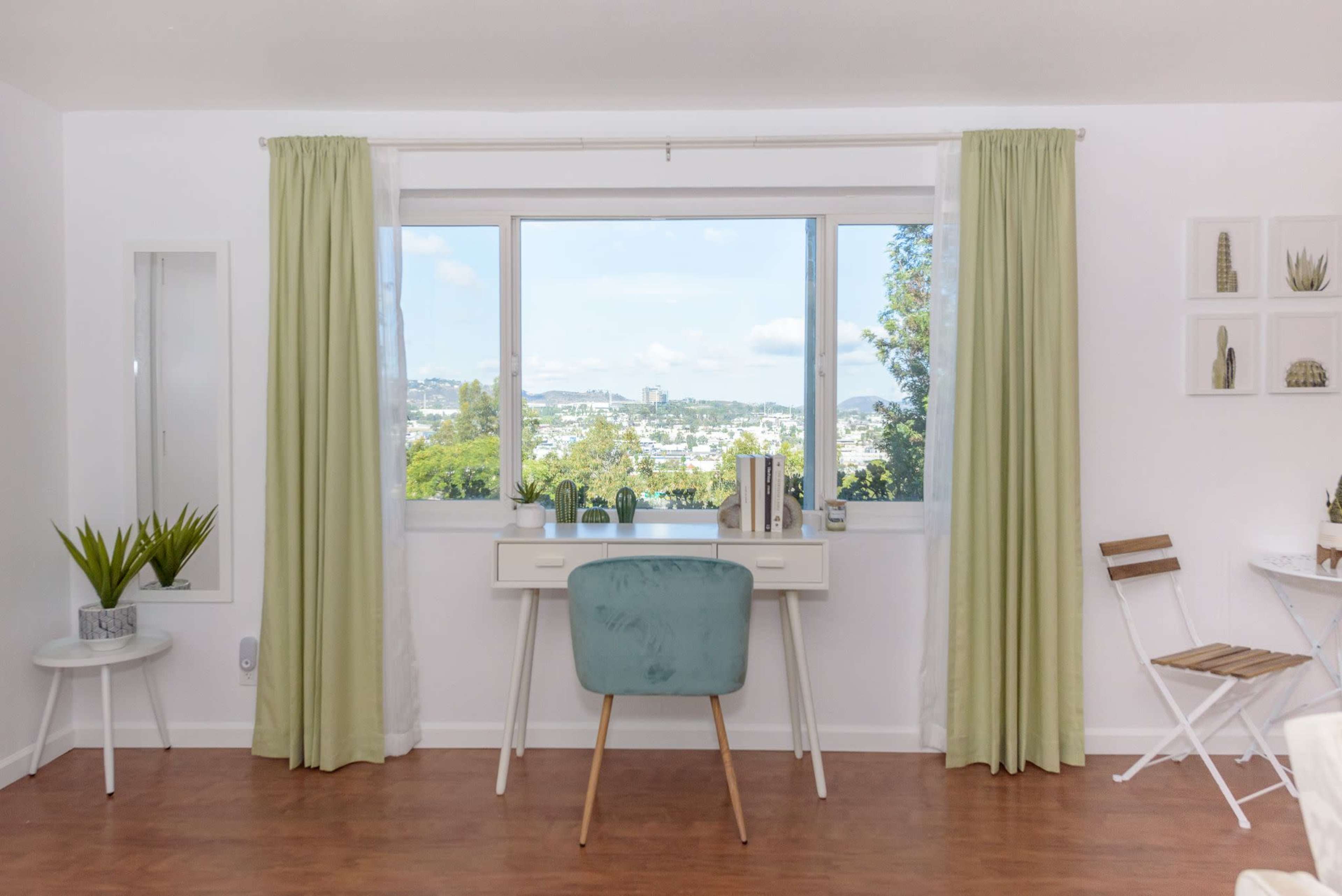 A bright room features a desk with a blue chair positioned near a window, framed by light green curtains, overlooking a city view.