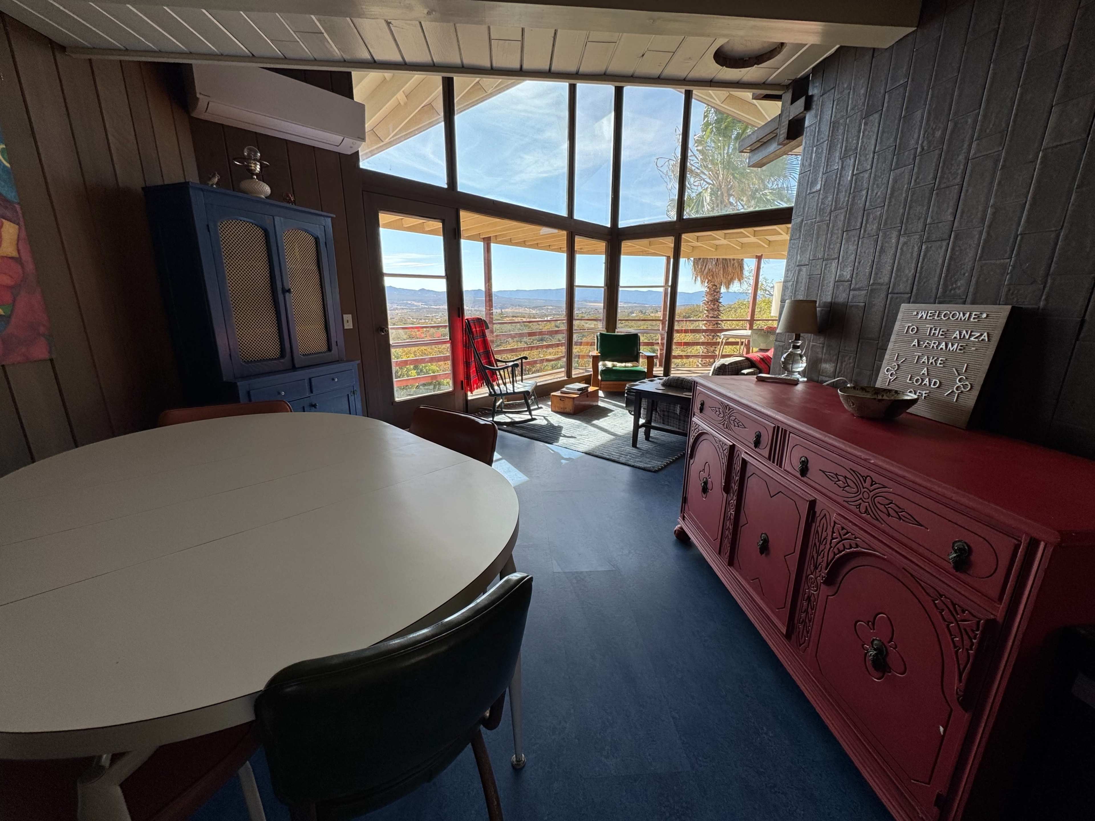 The image shows a well-lit interior space with a dining table, colorful furniture, and large windows offering a view of the outdoors.