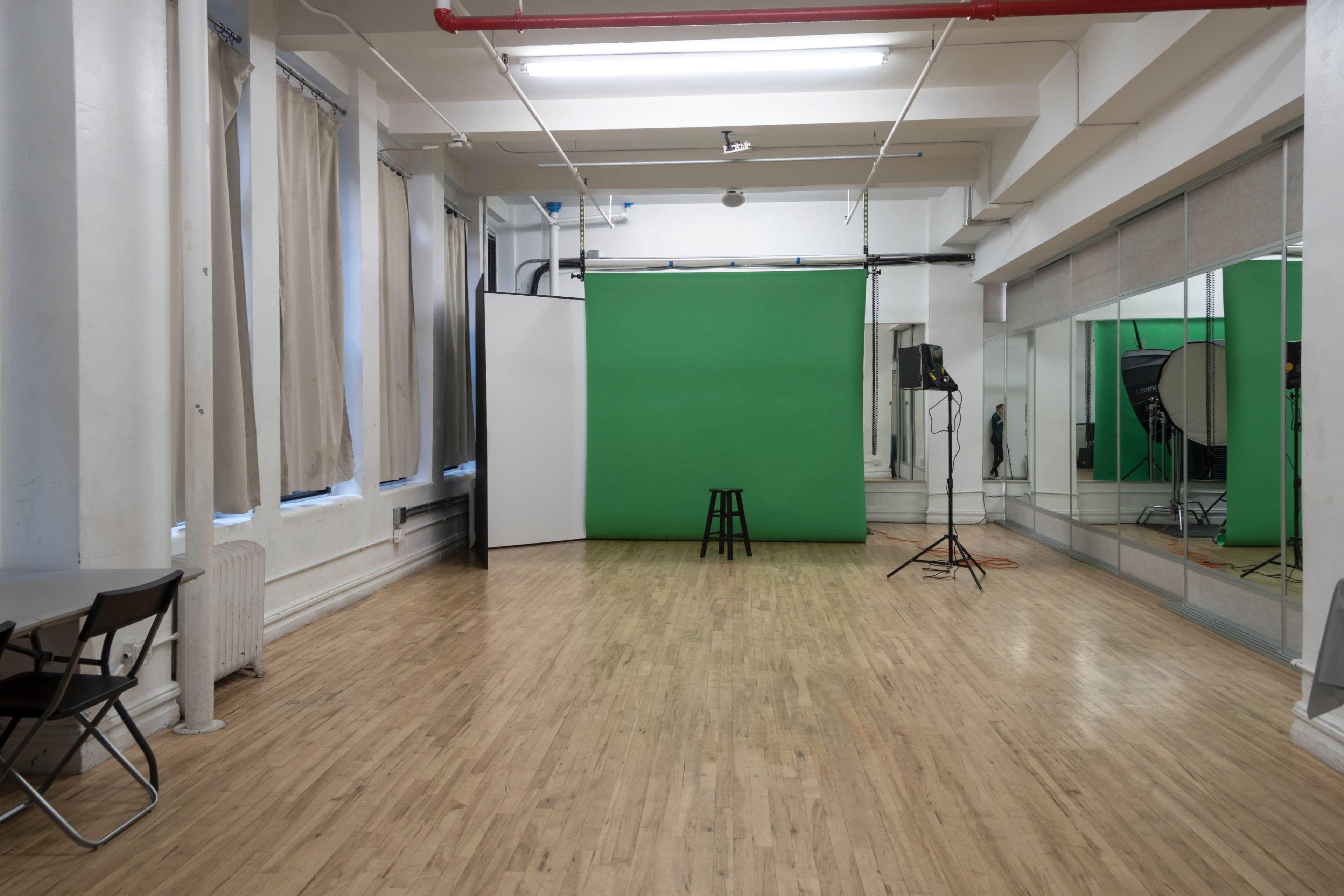 A spacious studio with a green backdrop, a stool, and various lighting equipment, surrounded by mirrors and natural light from large windows.