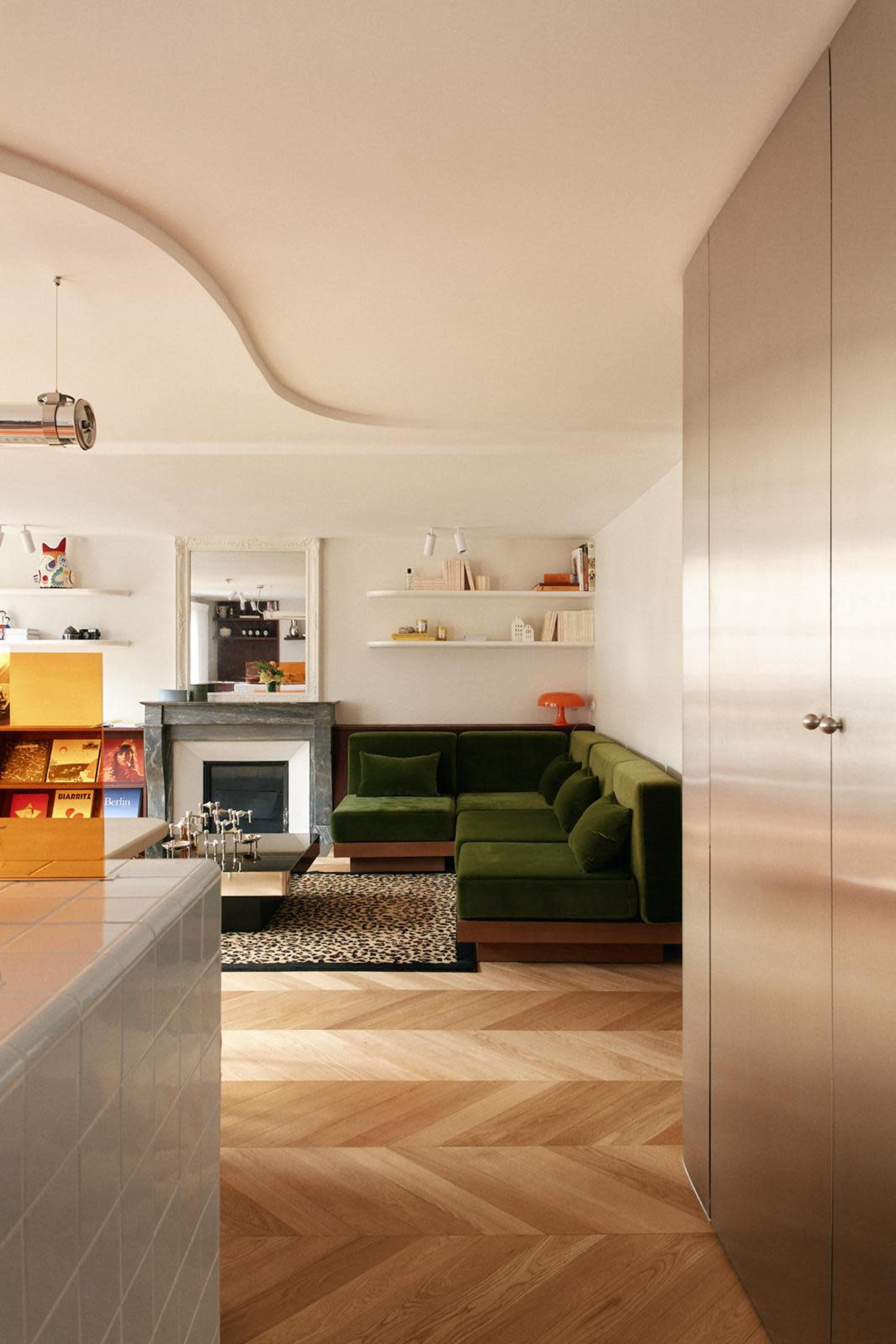 The image shows a modern living area featuring a green sectional sofa, a marble fireplace, and wooden floors with a herringbone pattern.