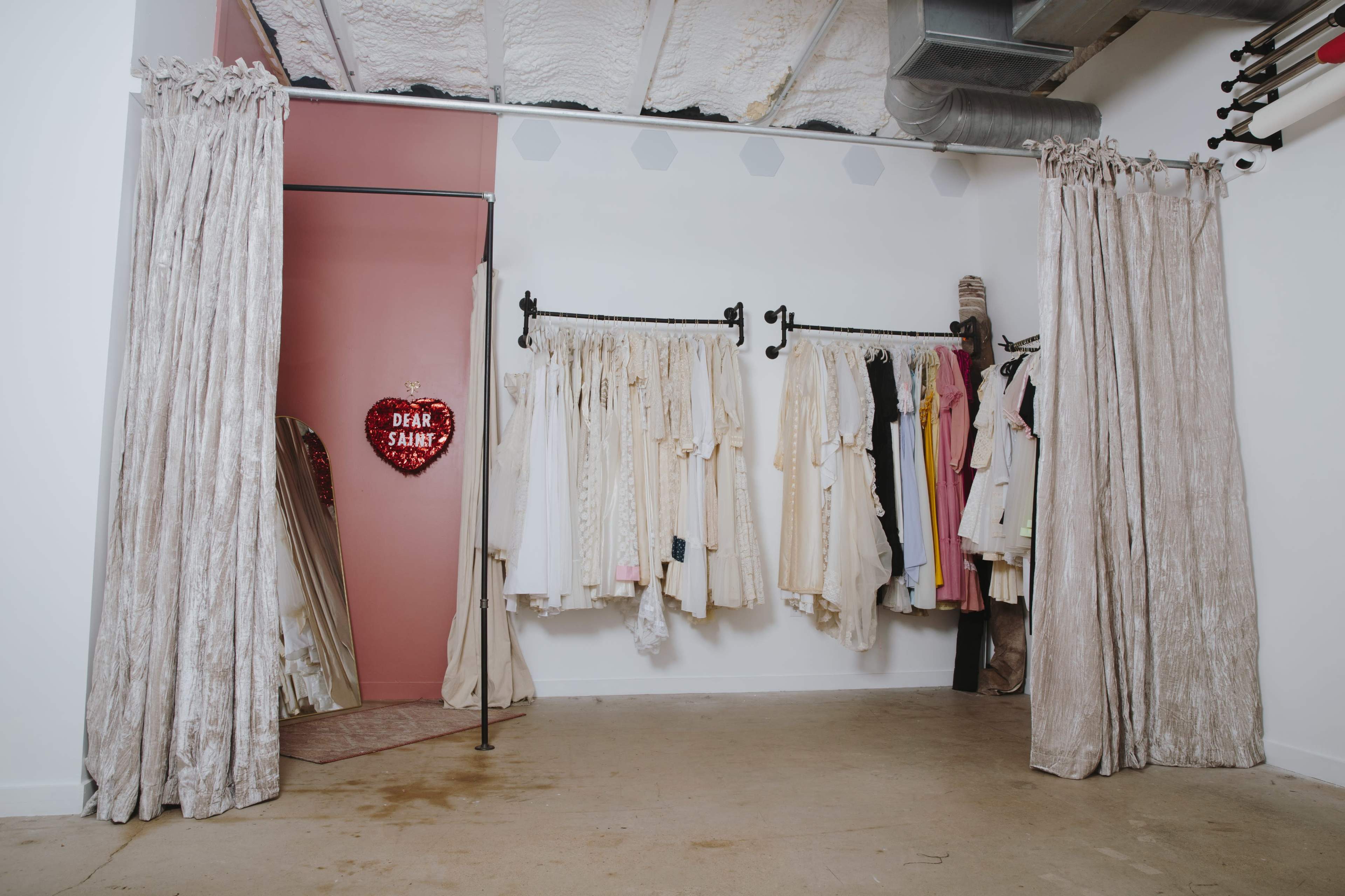 A clothing display area with hanging garments in light colors and a decorative heart-shaped sign on a pink wall.