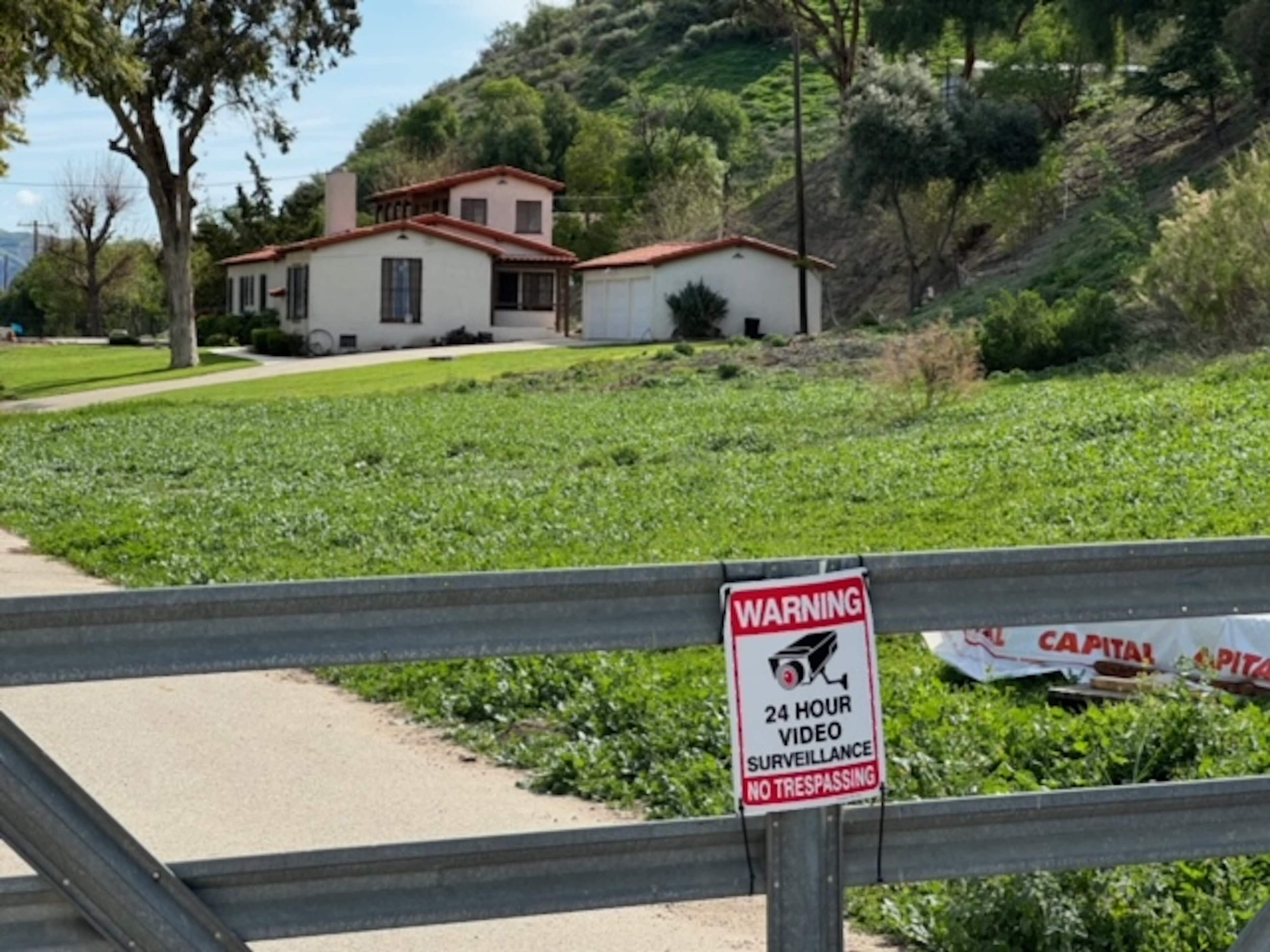 A house with a red-tiled roof is situated on a hillside, with a green field in front and a gated entrance marked by a warning sign about video surveillance.