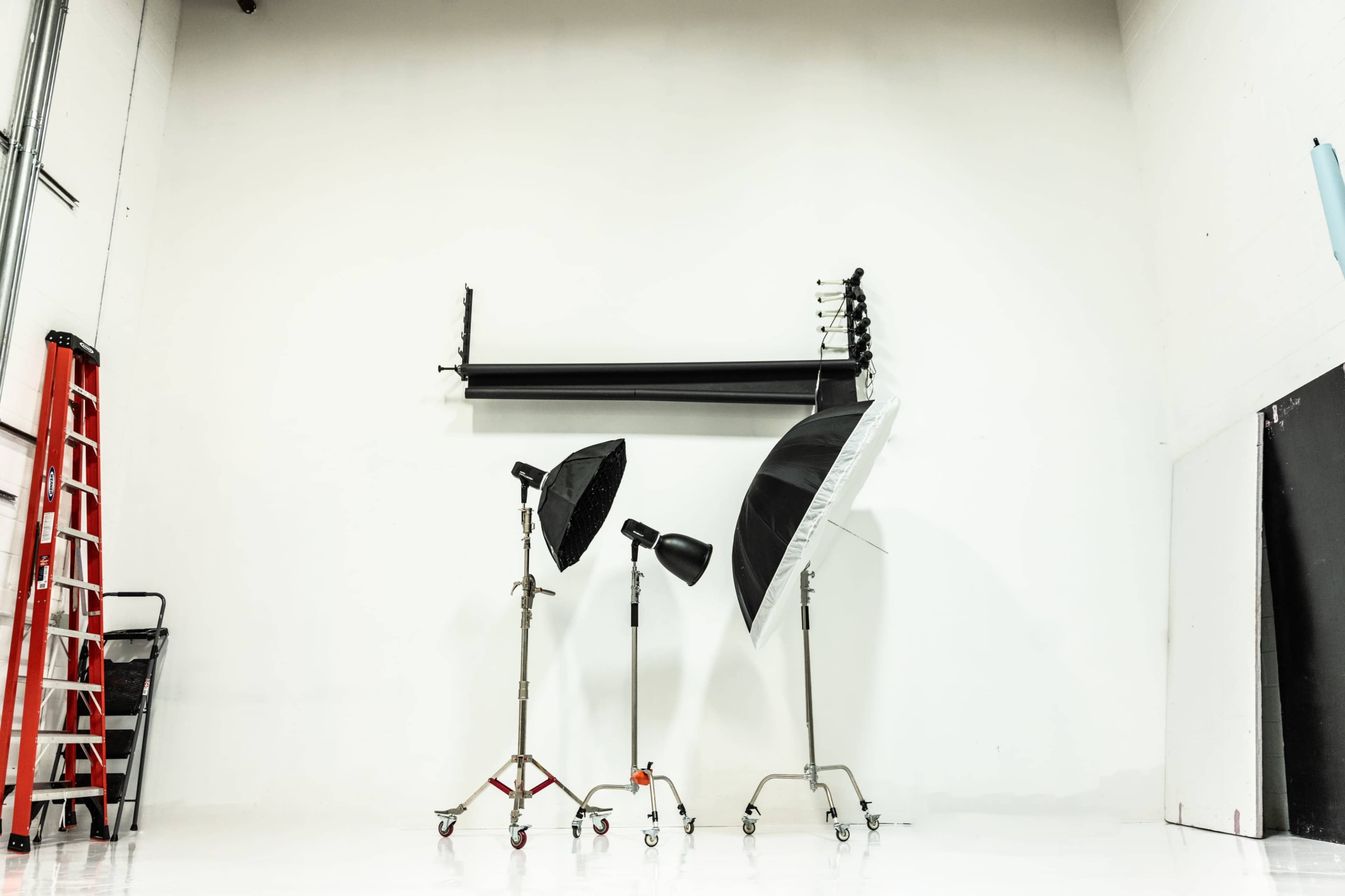 The image shows a photography studio with three light stands and a ladder against a blank white wall.