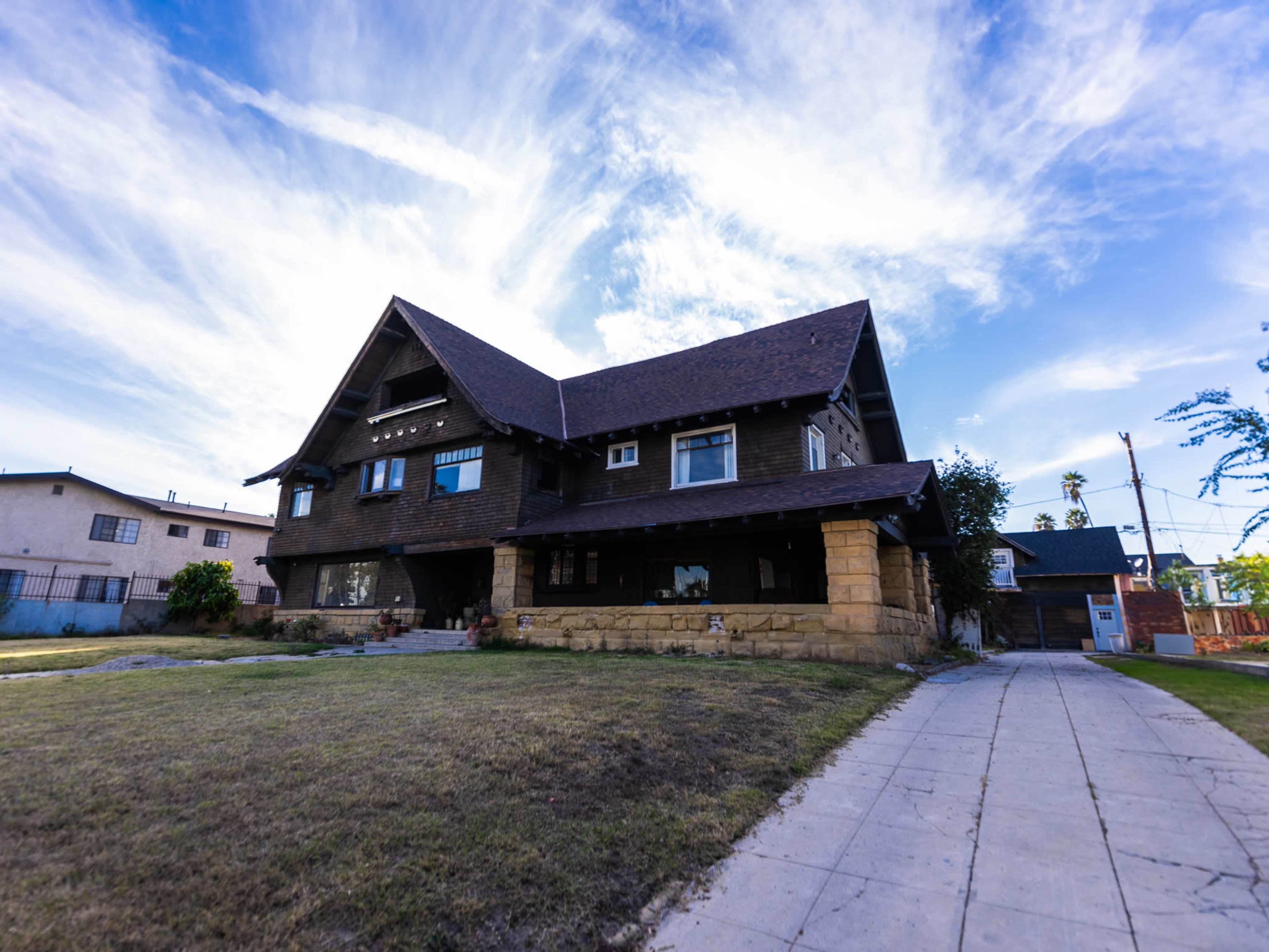 A large, two-story brown house with a steeply pitched roof and stone foundation is set against a blue sky with scattered clouds.