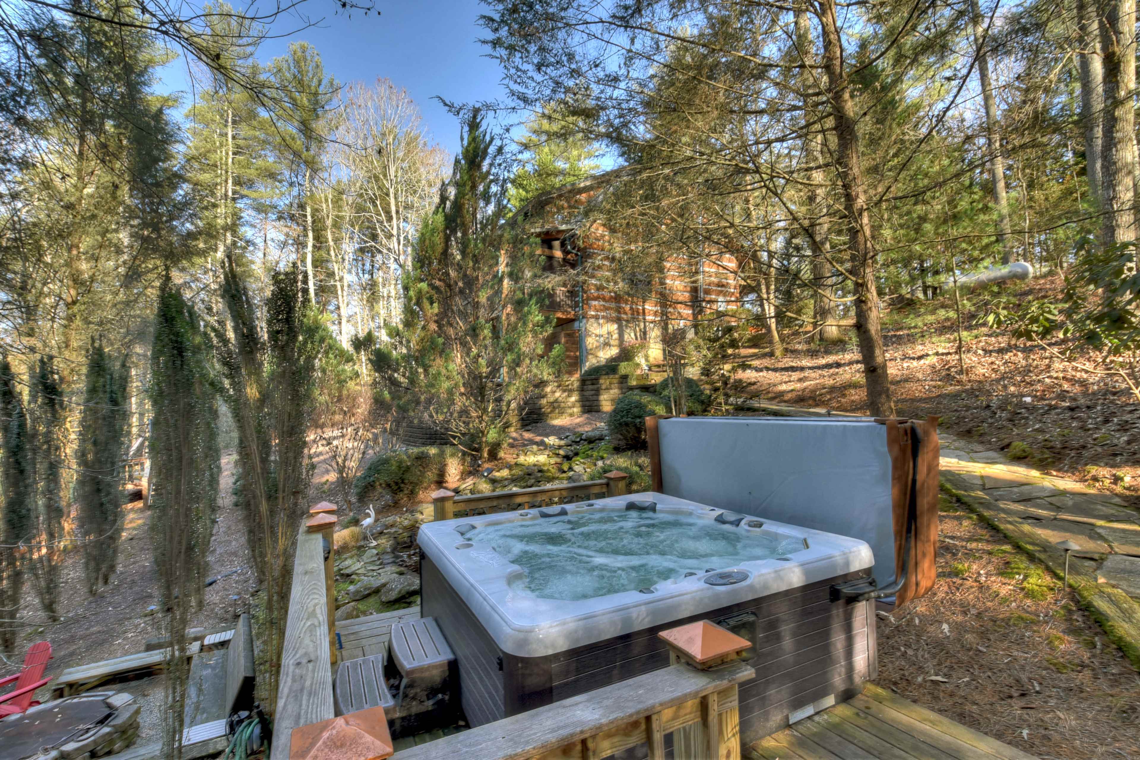 A hot tub sits on a wooden deck surrounded by trees and a pathway leading to a cabin.