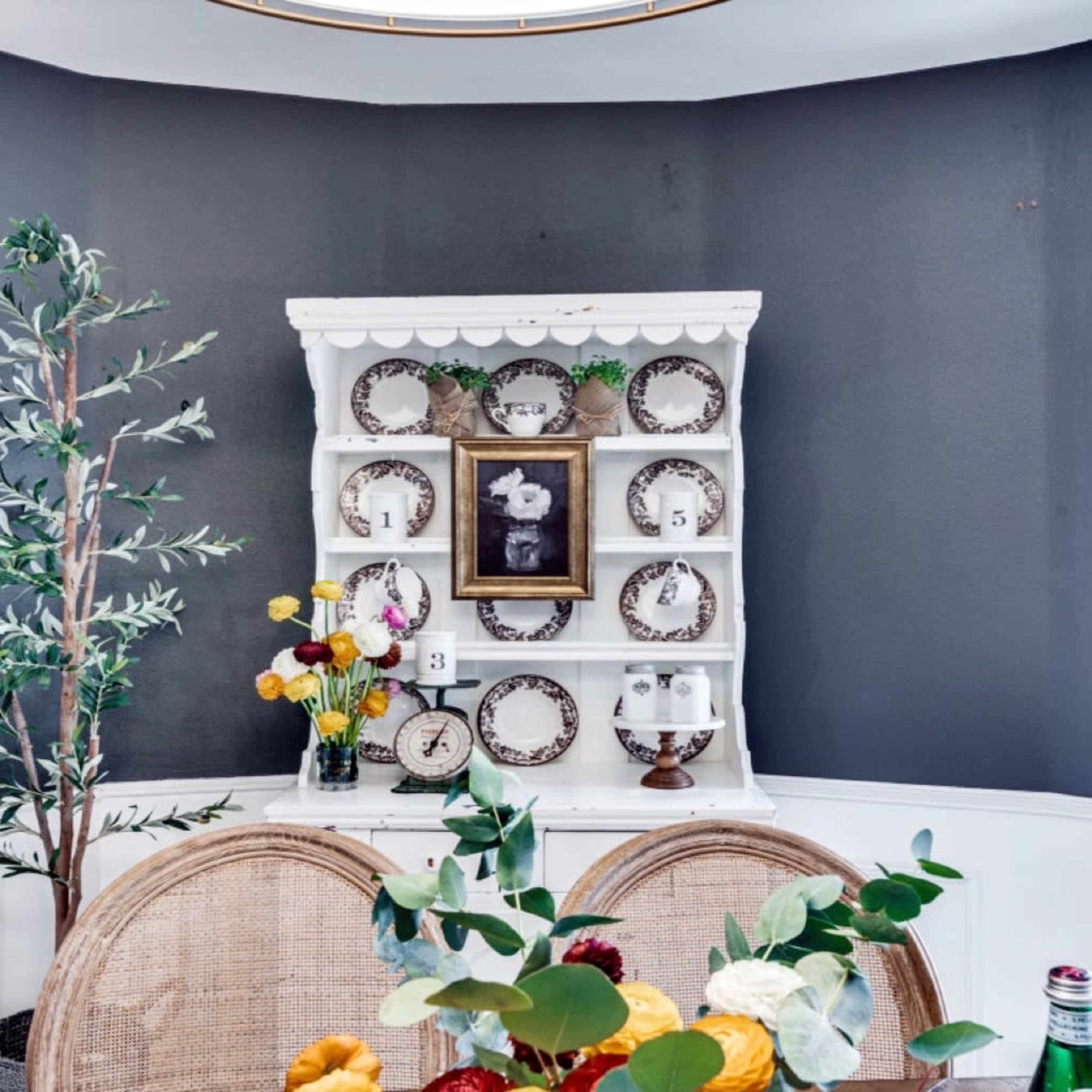 A white wooden hutch displays decorative plates and a framed artwork against a dark gray wall, complemented by a vase of flowers on the table in the foreground.