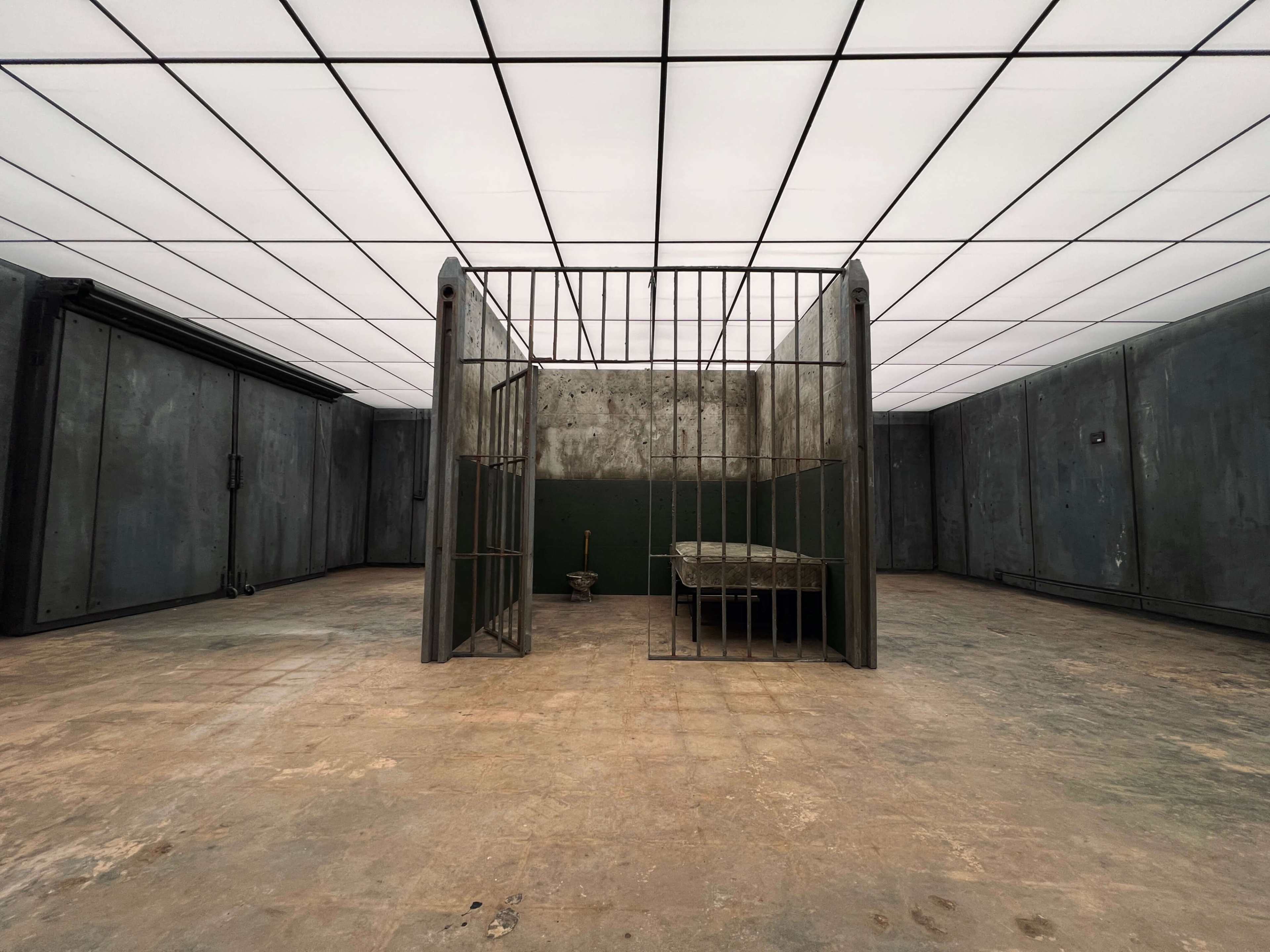 The image shows an empty, industrial-looking room with a metal cage in the center containing a concrete bench and a small object on the floor.