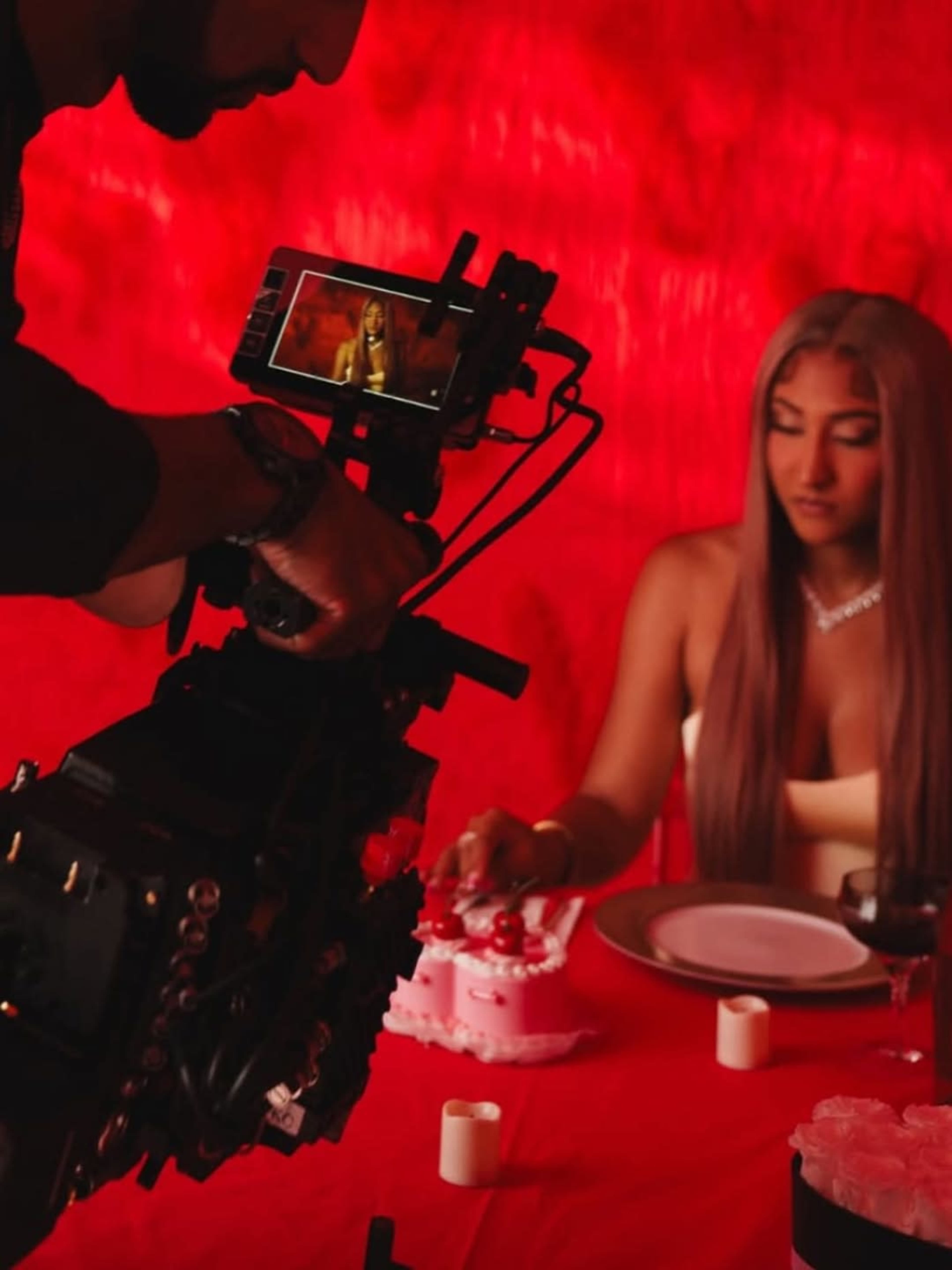 A camera operator captures a woman sitting at a table with a birthday cake against a red backdrop.