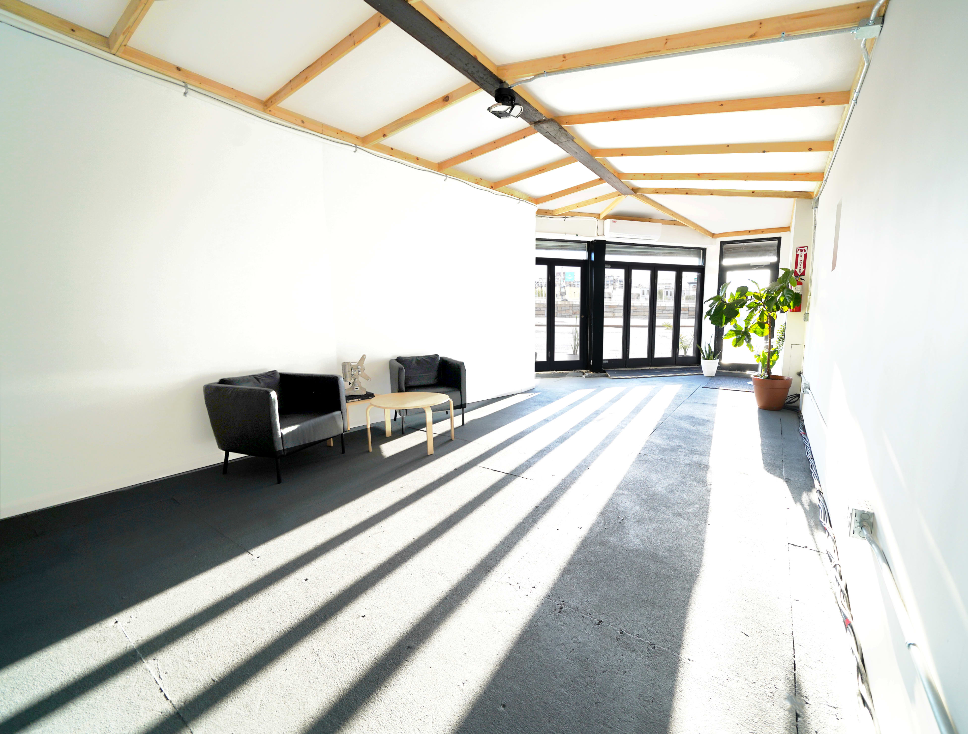 A bright, empty room with a wooden beam ceiling, a couple of black chairs, a small table, and a large potted plant near the entrance.