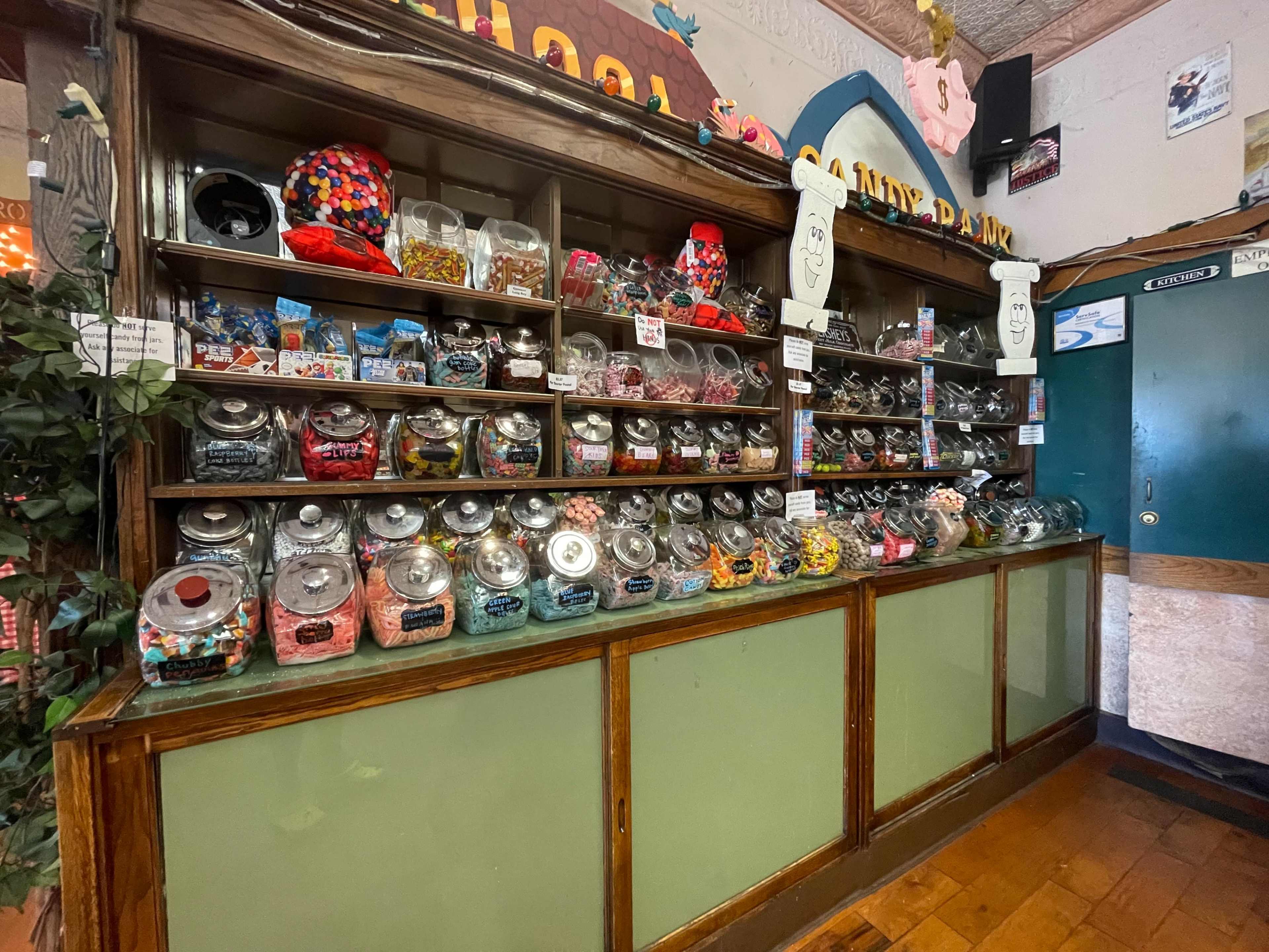 A wooden candy display with multiple glass jars filled with various colorful candies, set against a wall adorned with decorations.