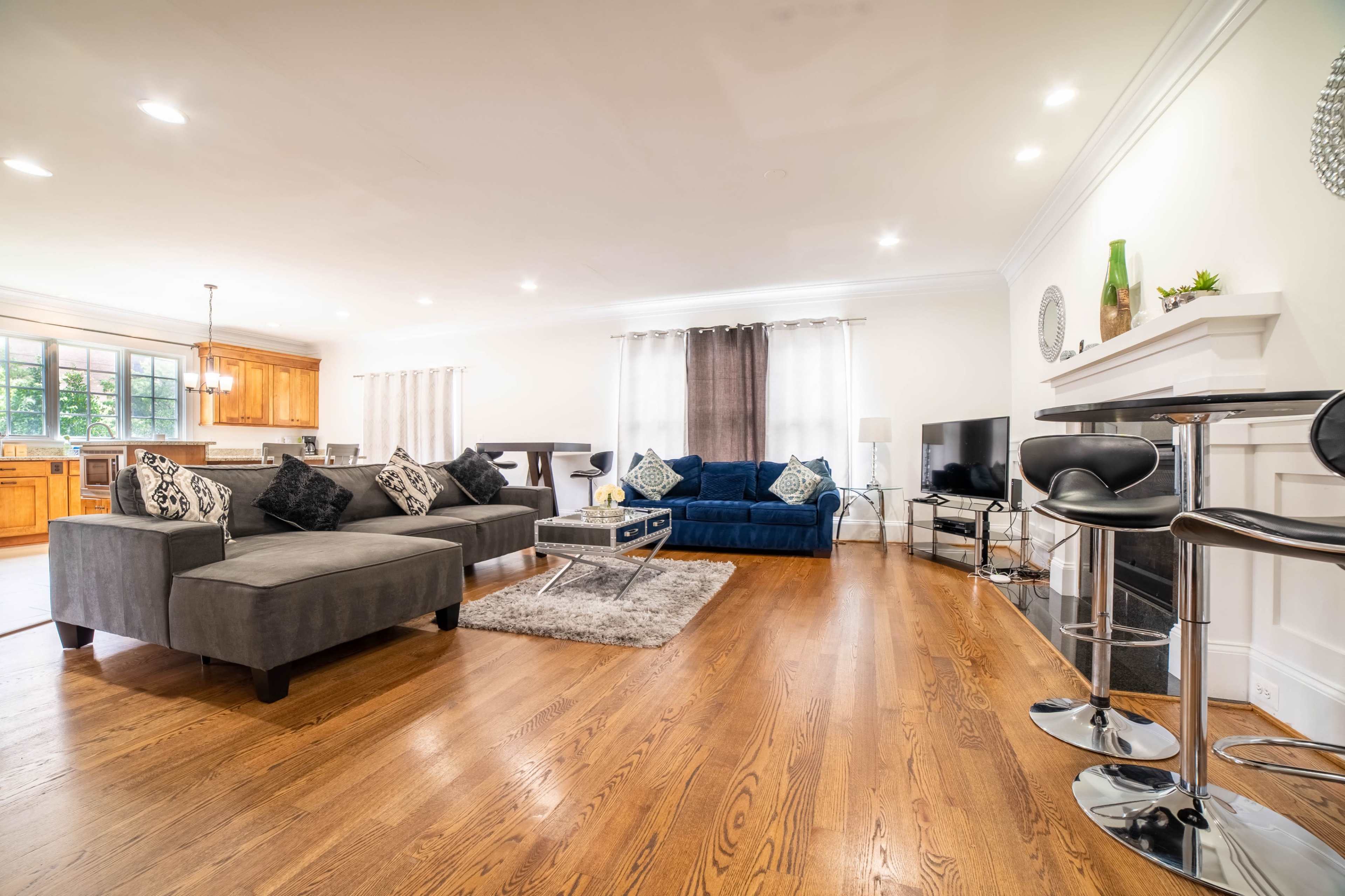 The living room features a blue sofa, a gray sectional, a coffee table, and a television, with wooden flooring and a kitchen visible in the background.