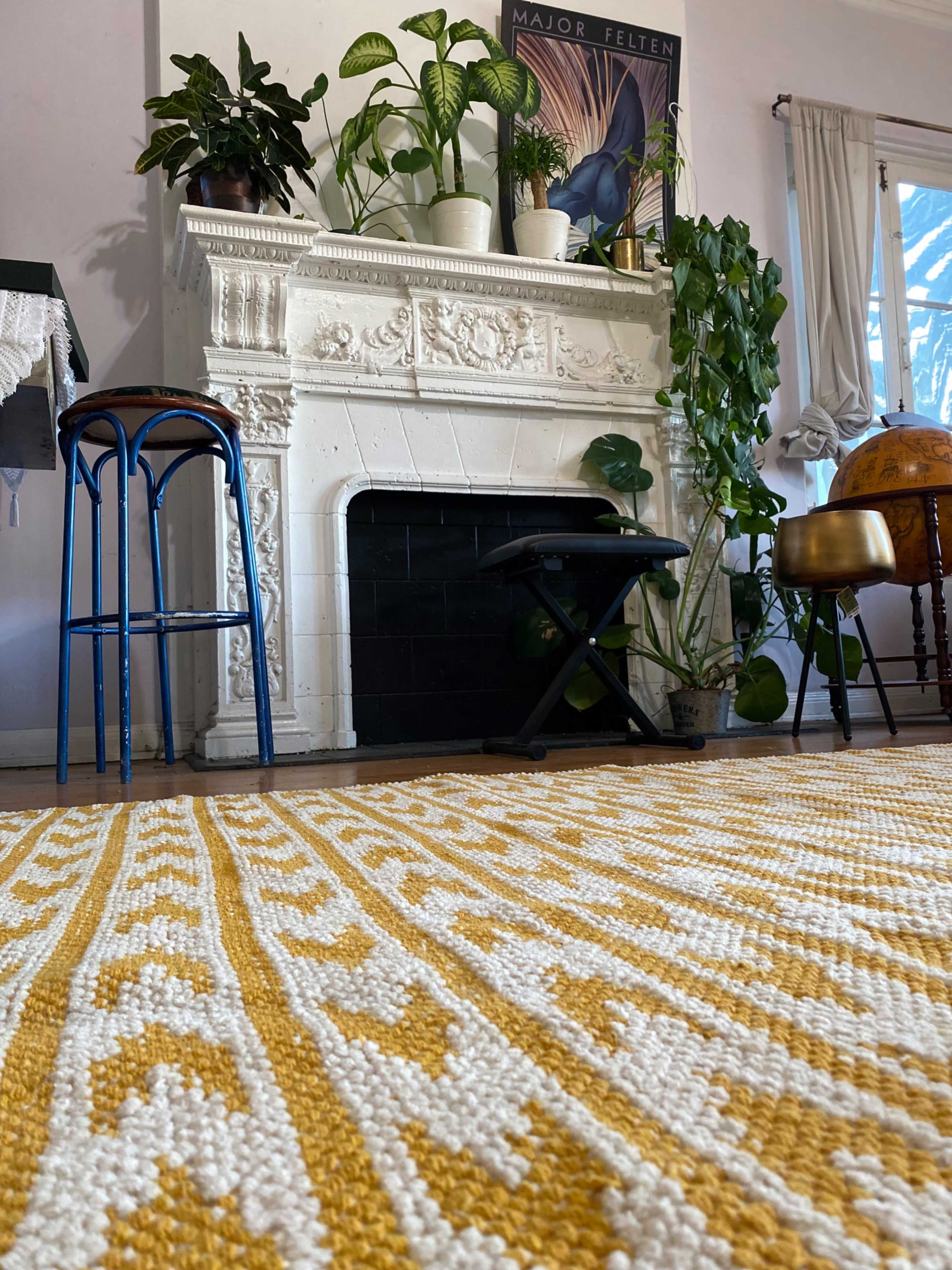 The image shows a living room with a white fireplace surrounded by plants, a patterned yellow and white rug on the floor, and various furniture pieces, including stools and a globe.