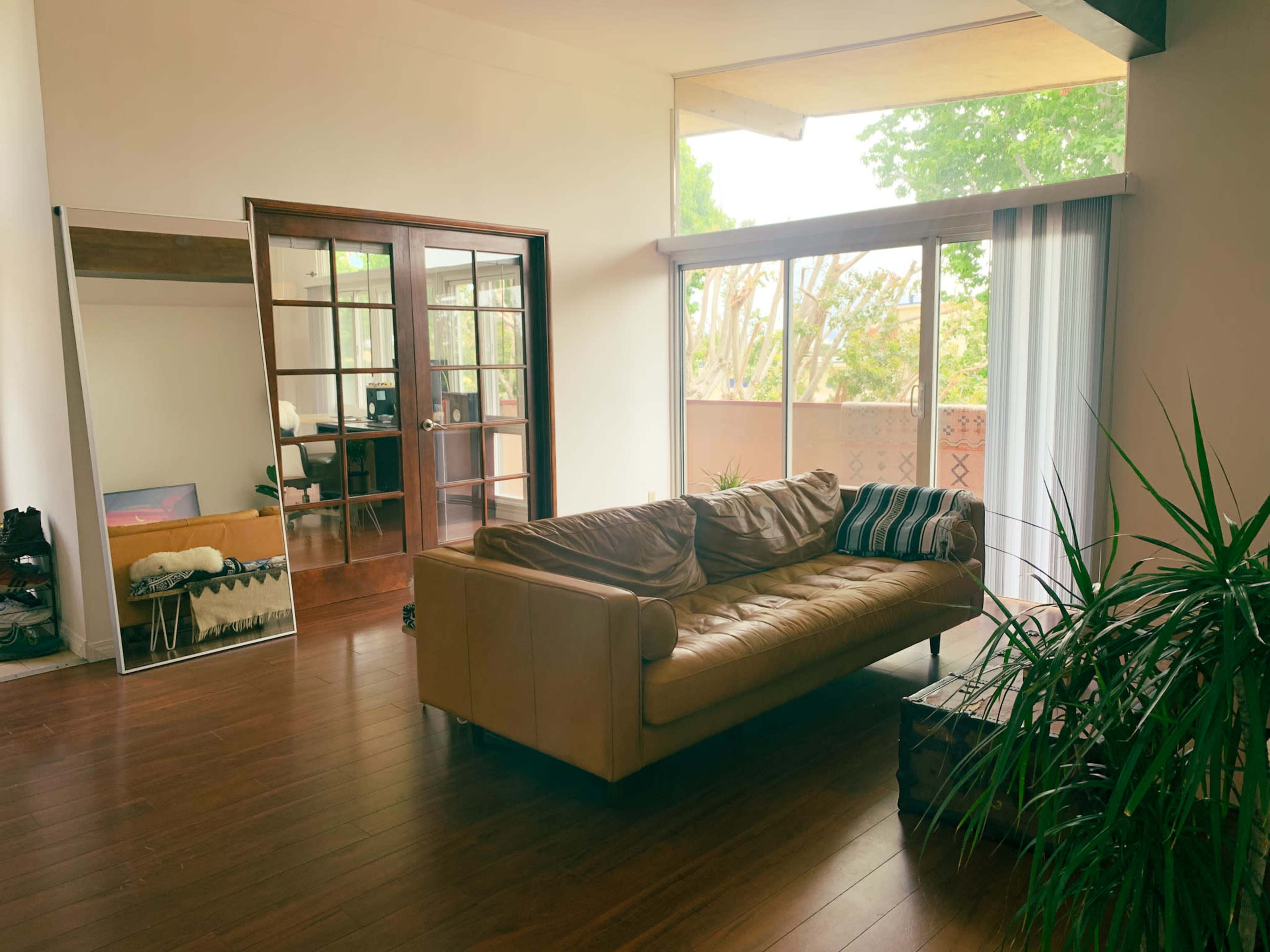 A spacious living room with a leather sofa, a large mirror, and glass doors leading to a balcony surrounded by greenery.
