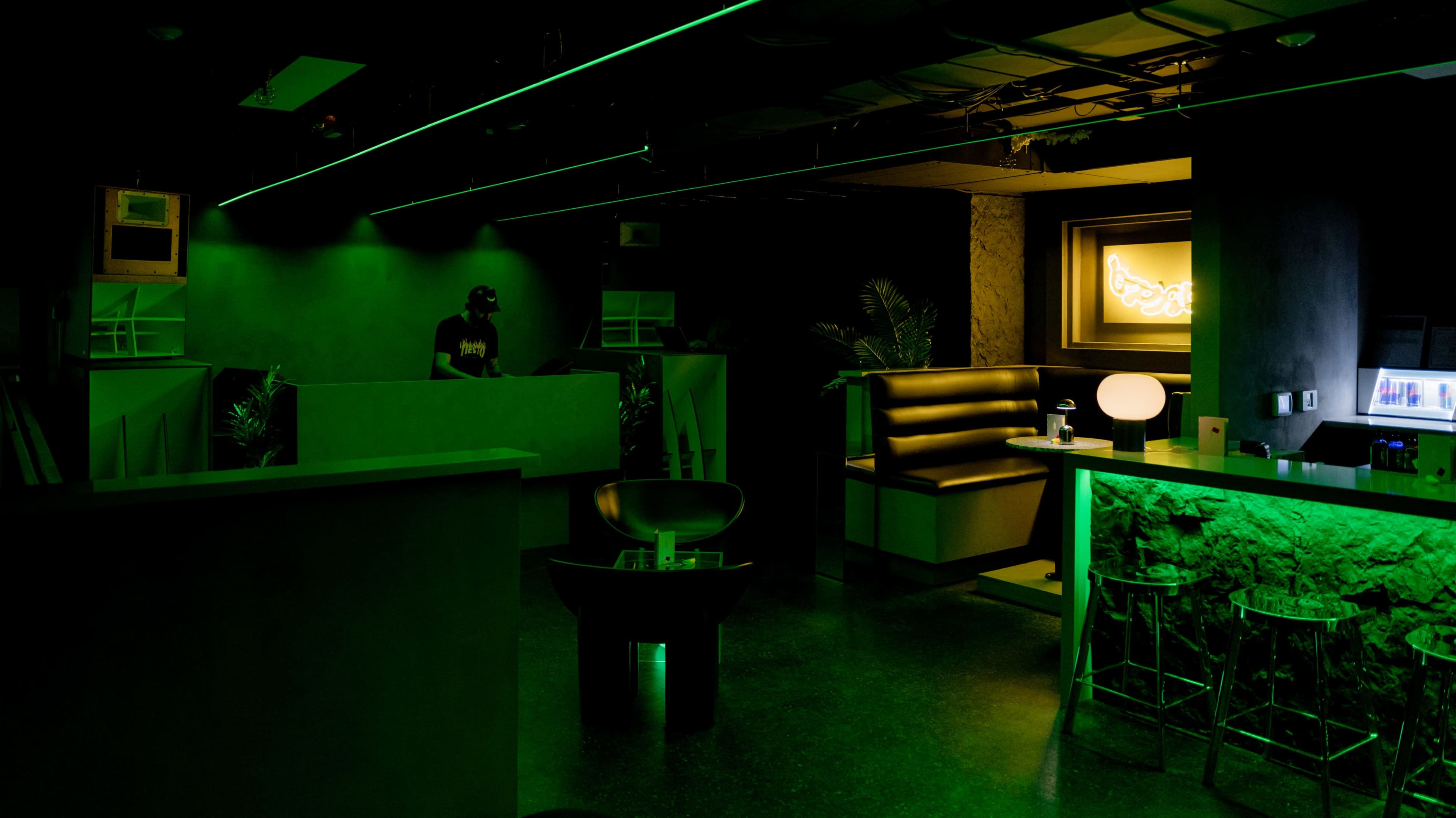 A dimly lit lounge features green lighting, a bar with stools, and a DJ at the back.