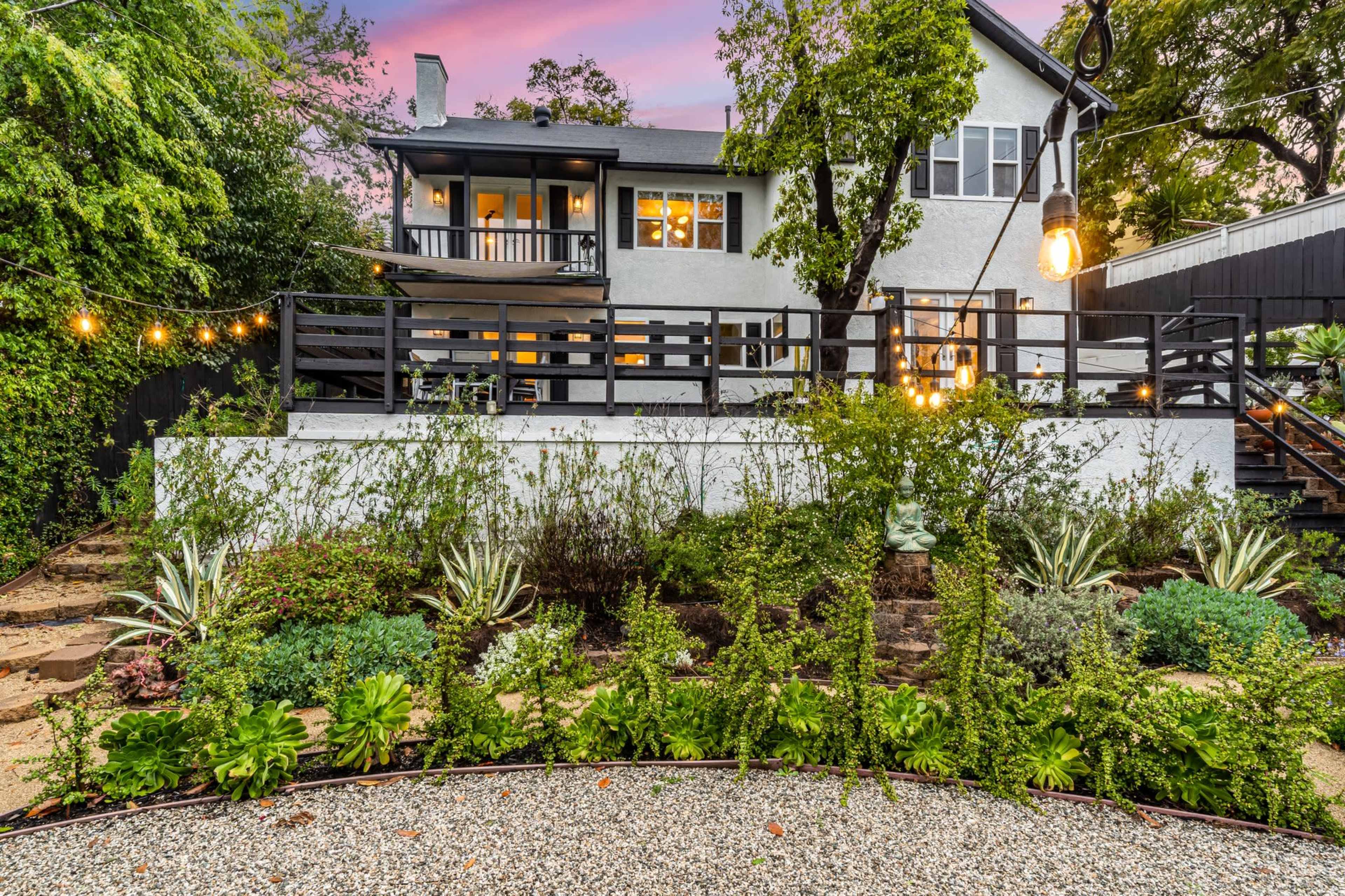 A well-kept garden with various plants and a circular stone pathway leads to a modern two-story house with a deck, surrounded by trees and illuminated by string lights.